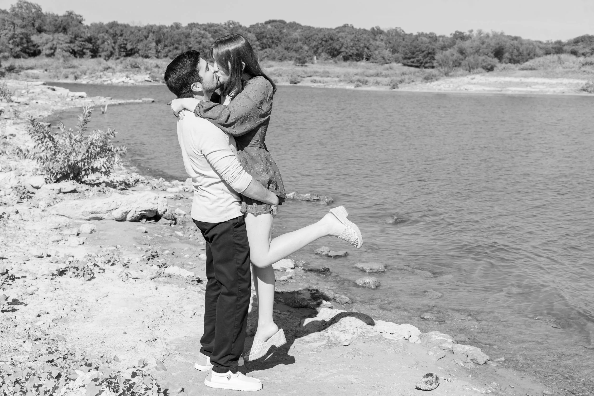 A couple kissing by a lake with the woman lifting her foot off the ground.