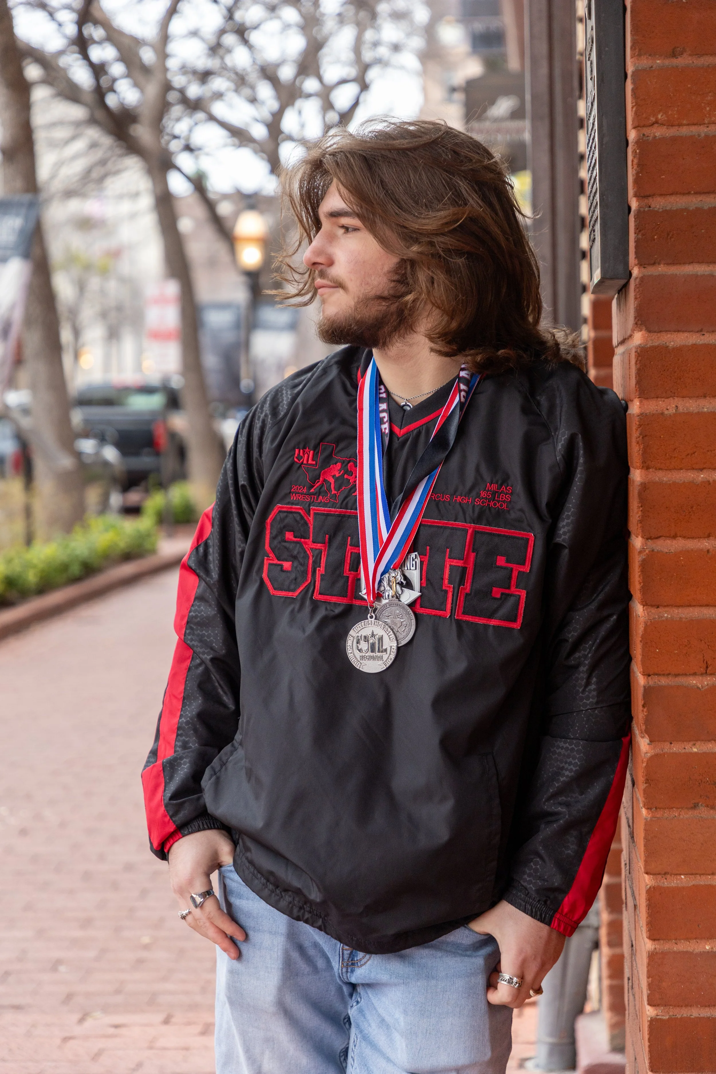 A young man with long brown hair and a beard leaning against a brick wall, wearing a black and red athletic jacket with medals around his neck, outdoors on a sidewalk with trees and parked cars in the background.
