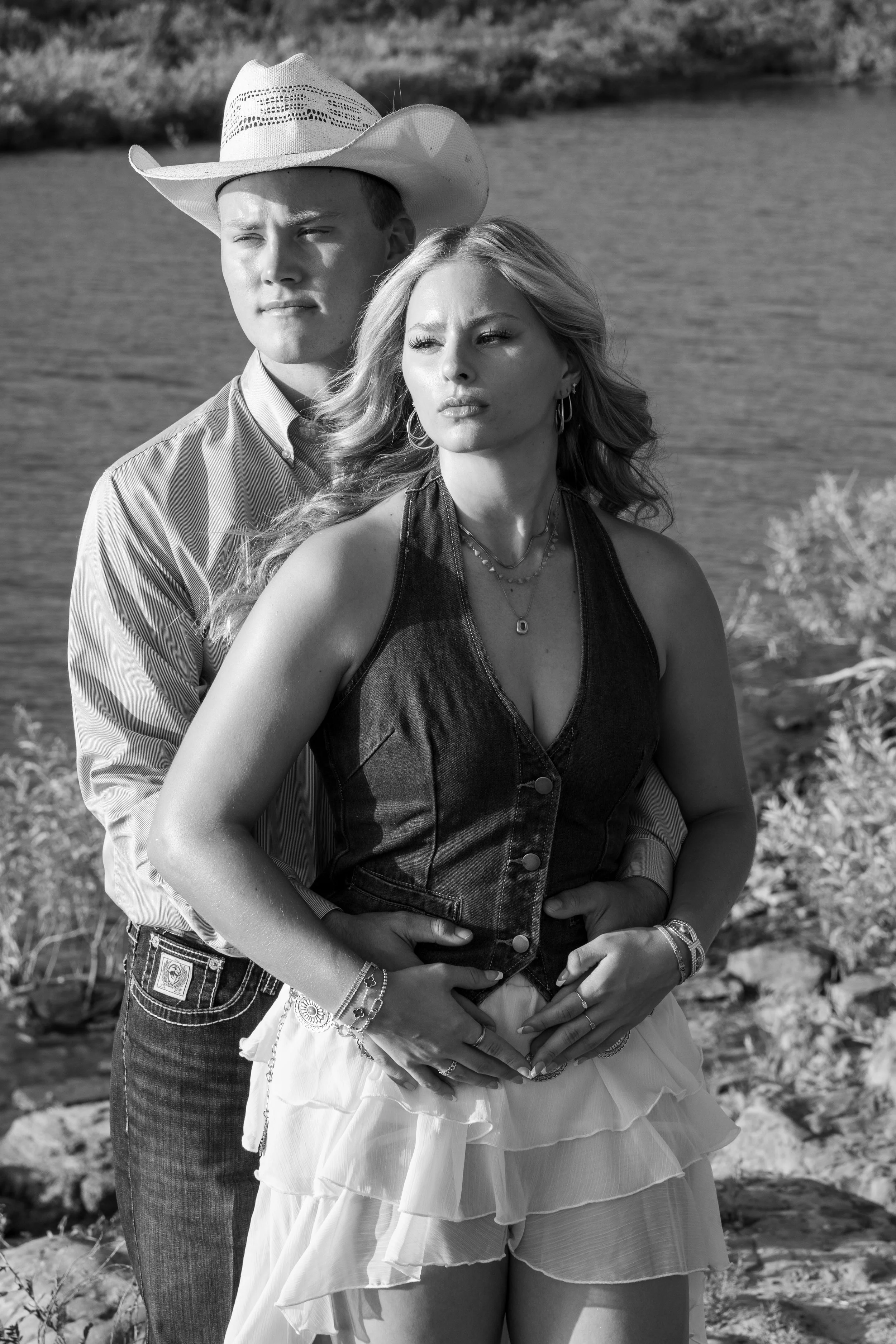 A black and white photo of a young man wearing a cowboy hat and a young woman with wavy hair, multiple necklaces, and bracelets, standing outdoors near a body of water, both gazing into the distance.