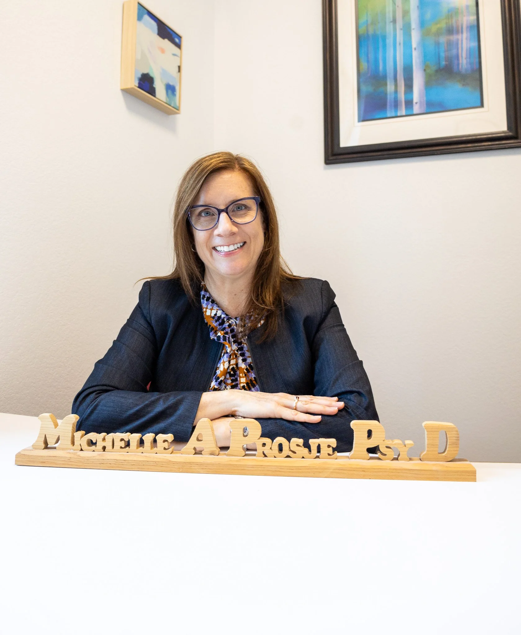 A woman with shoulder-length brown hair and glasses, smiling, sitting at a white table with her hands folded. In front of her are wooden letters spelling "Michele A. Rosé, Psy. D." She is wearing a dark blazer and a patterned blouse. The background i