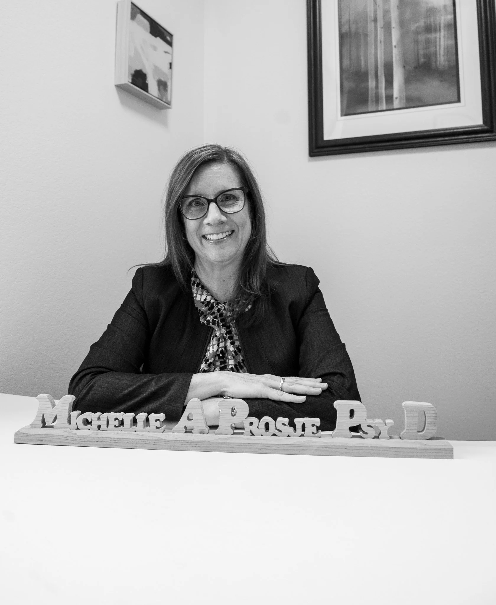 A woman wearing glasses and a blazer smiling at the camera, sitting at a table with a nameplate that reads "Michelle A. Rose, Ph.D." in front of her, with framed art on the wall behind her.