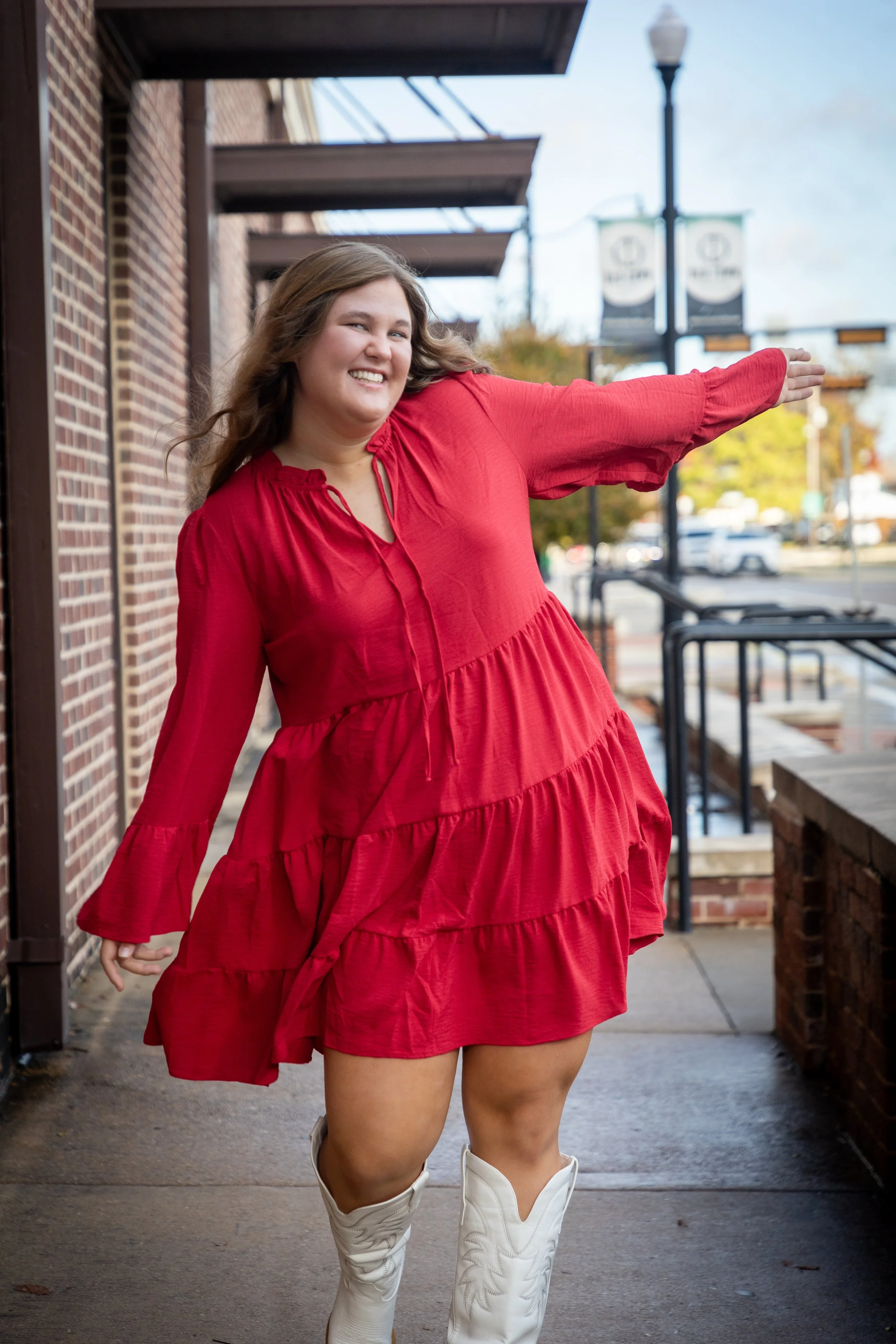 A smiling woman in a red dress and white cowboy boots posing outdoors on a sidewalk.