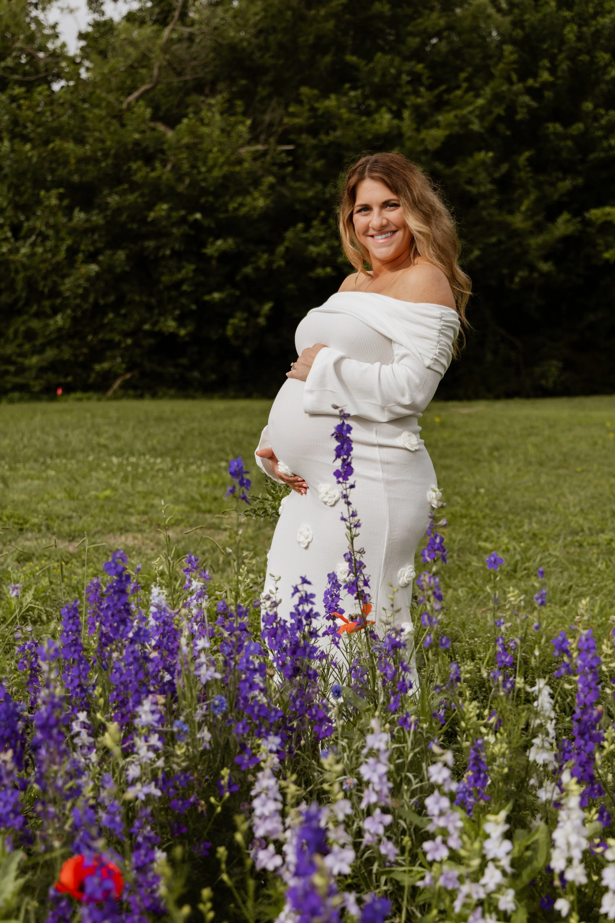 A pregnant woman smiling outdoors, standing among purple and red flowers with a grassy field and green trees in the background.