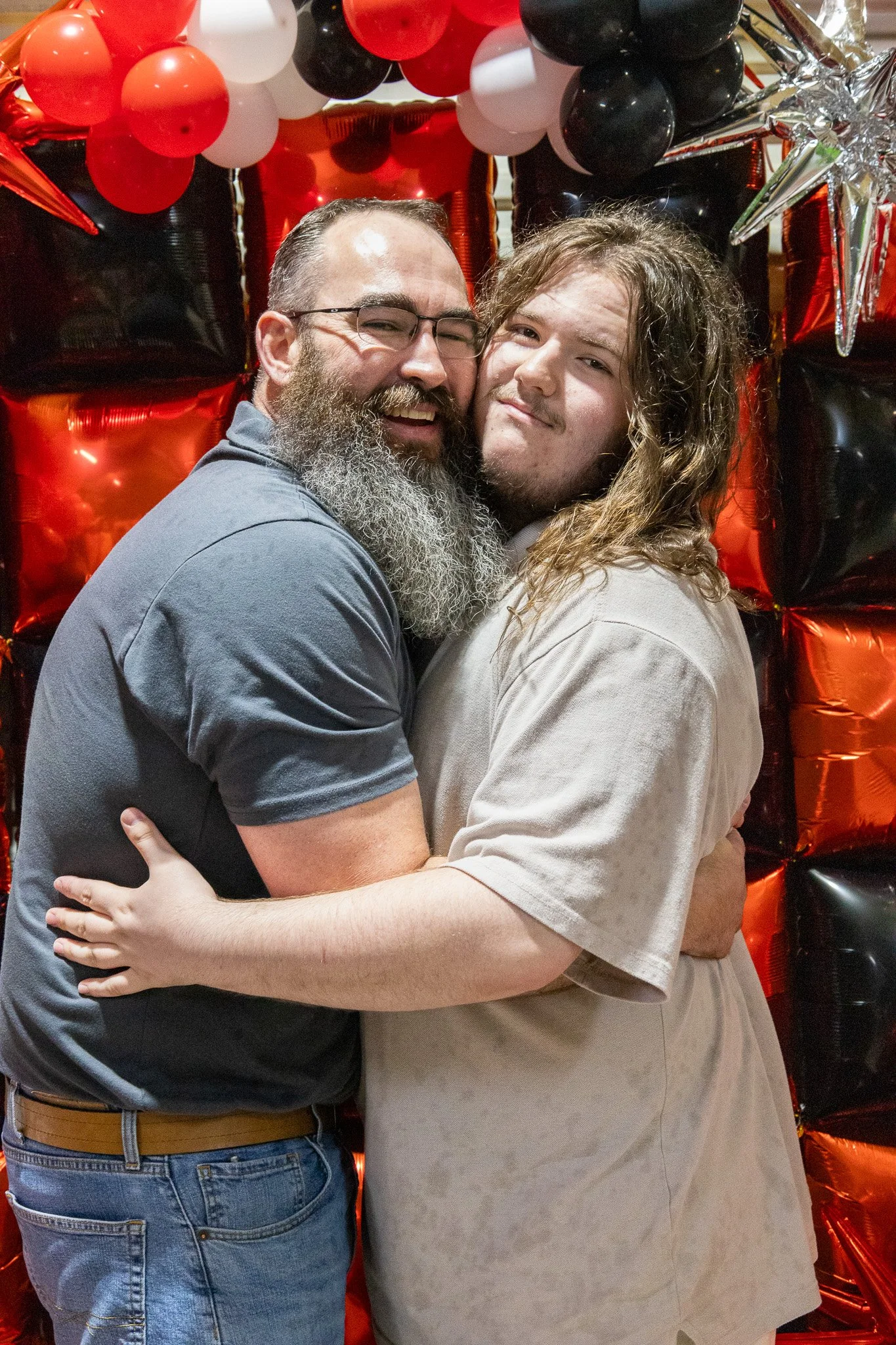 Two men hugging and smiling, standing in front of a backdrop made of red, black, white, and silver balloons.