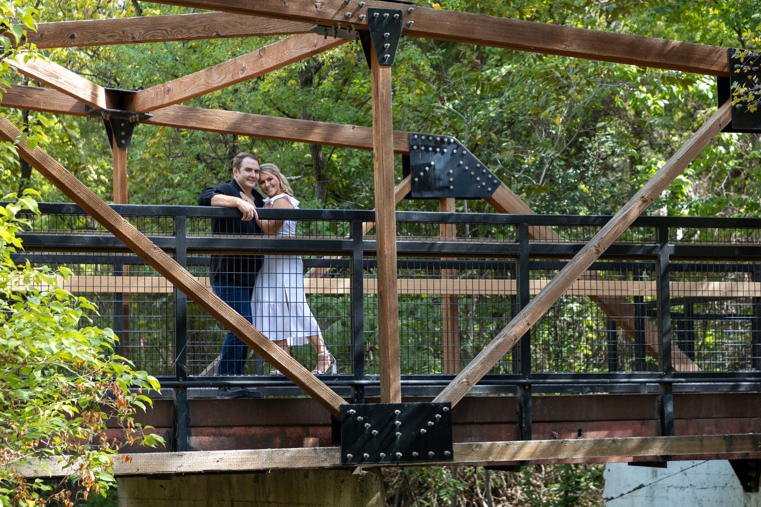 A cheerful couple standing on a wooden and metal bridge surrounded by lush green trees.