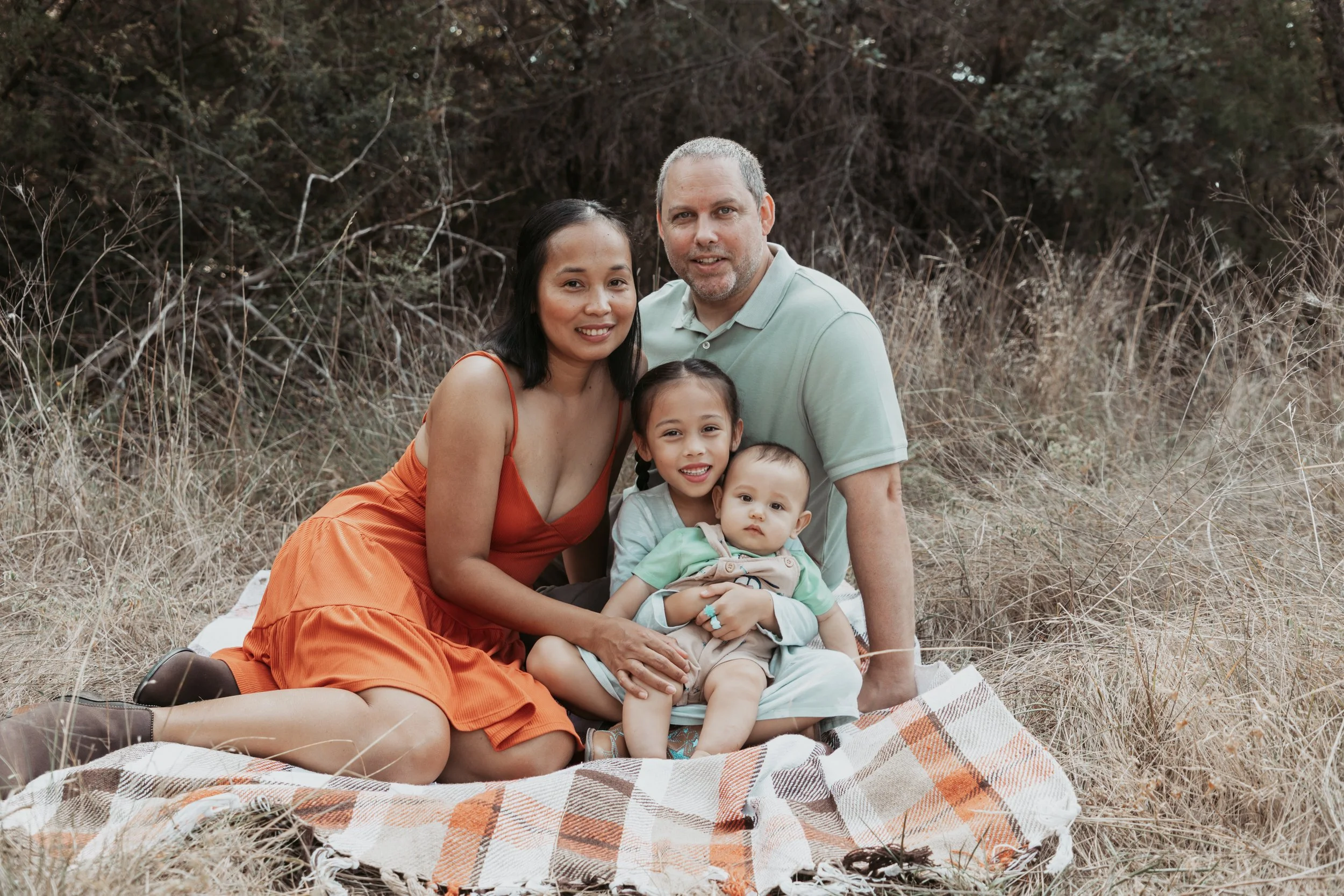 Family of four sitting on a blanket in a field with tall grass and trees in the background. The mother wears an orange dress, the father a light green polo shirt, the older daughter has braids and a light-colored shirt, and the younger baby wears a t