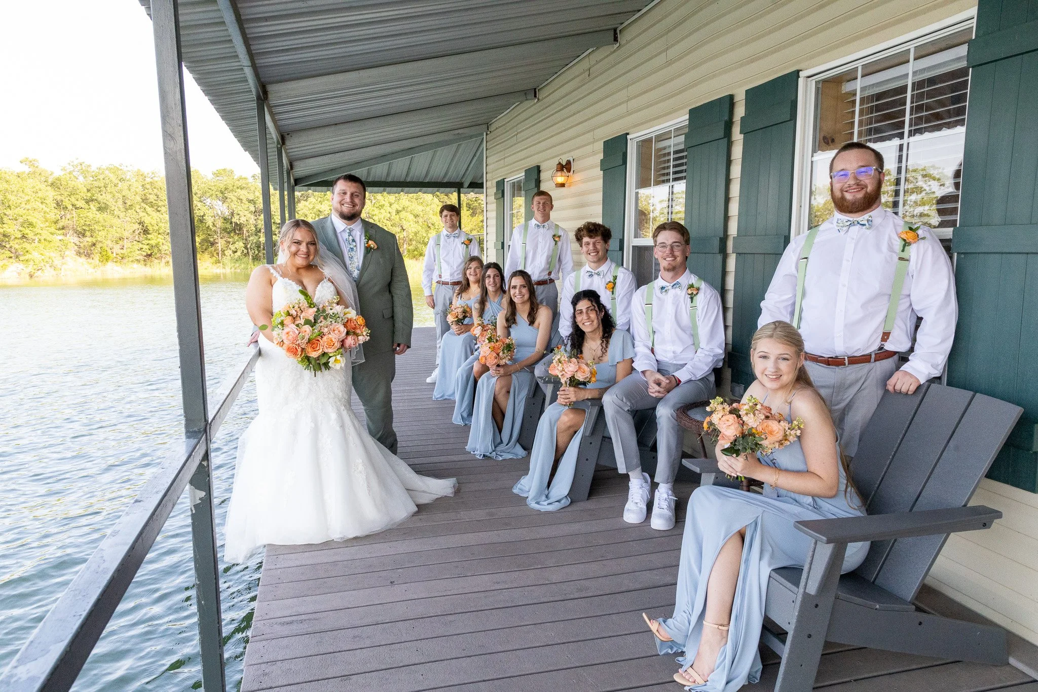 A group of wedding party members and a bride on a wooden deck by the water. The bride wears a white wedding dress and holds a bouquet. The groom and groomsmen wear light gray suits with colorful boutonnières and suspenders. The bridesmaids wear light