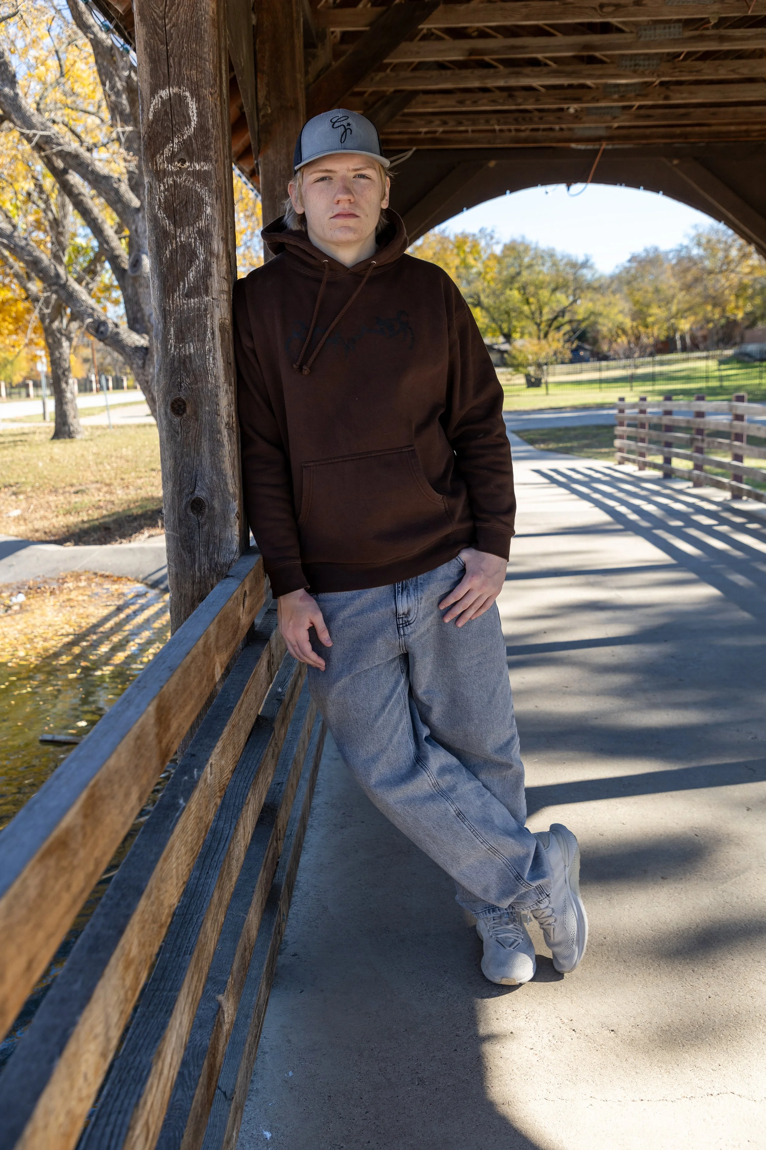 A young man leaning against a wooden support on a small bridge in a park, wearing a brown hoodie, light gray baggy jeans, white sneakers, and a gray cap, with trees and a clear blue sky in the background.