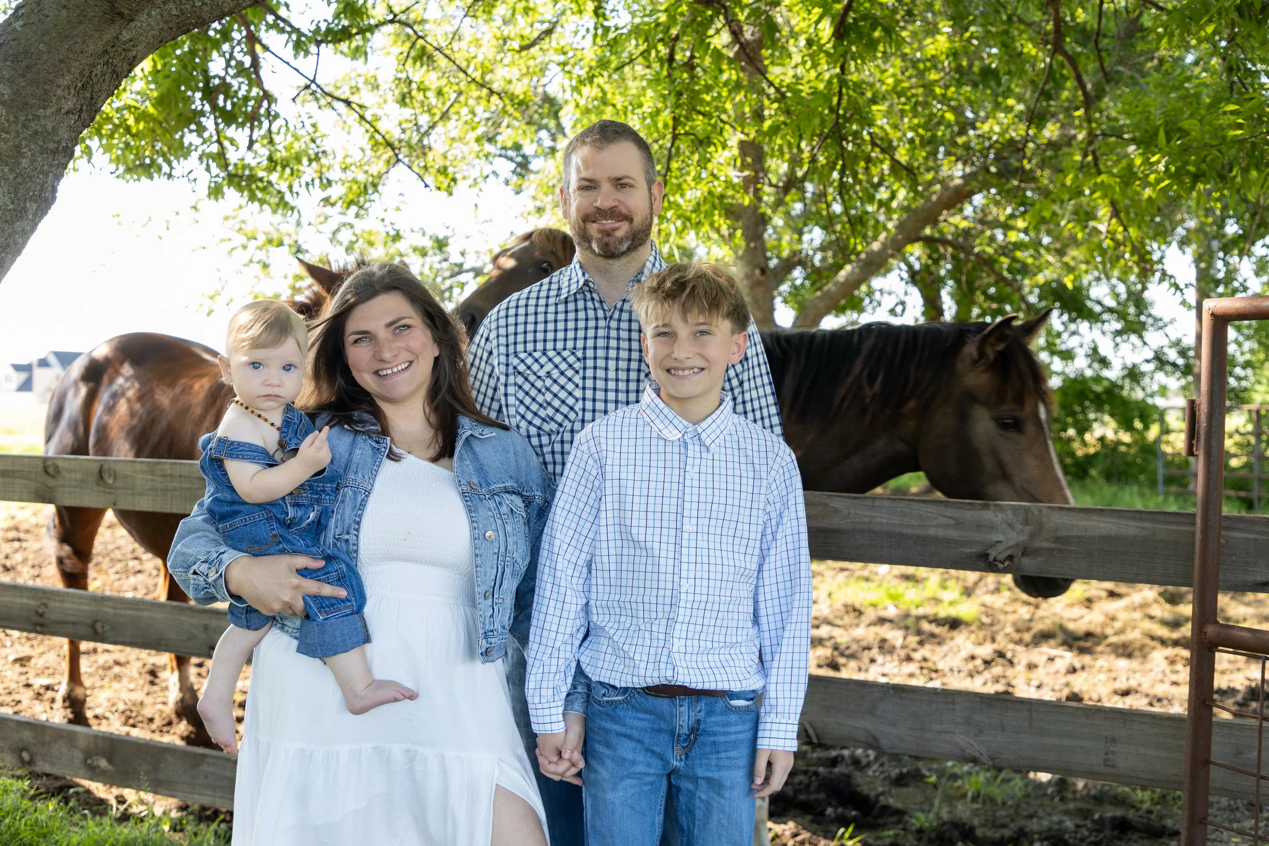 A family of four standing outdoors near a wooden fence with horses behind them, under a large tree with green leaves, during daytime.