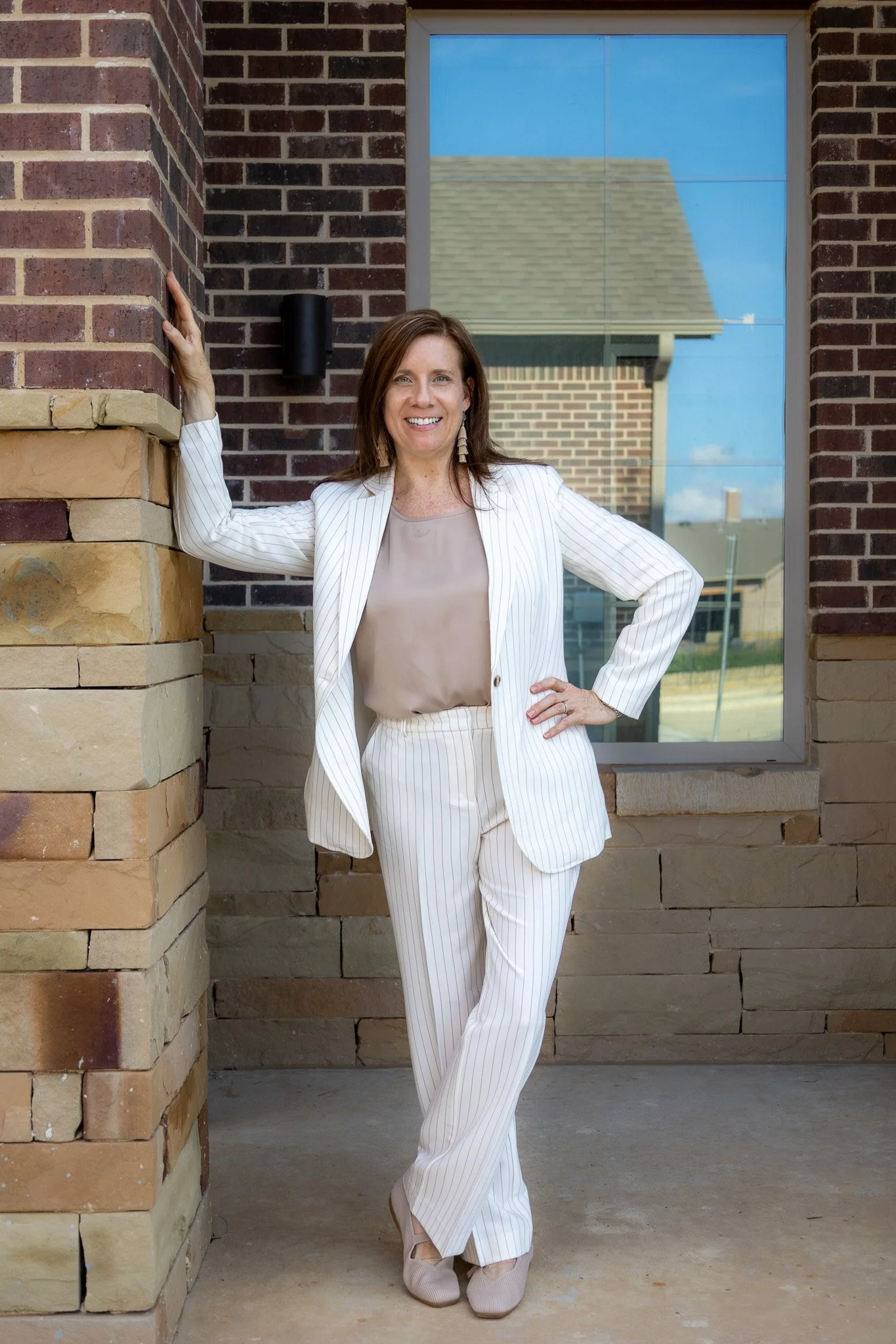 A woman in a white pinstripe suit standing outside a brick building, smiling with one arm raised and resting on the wall.