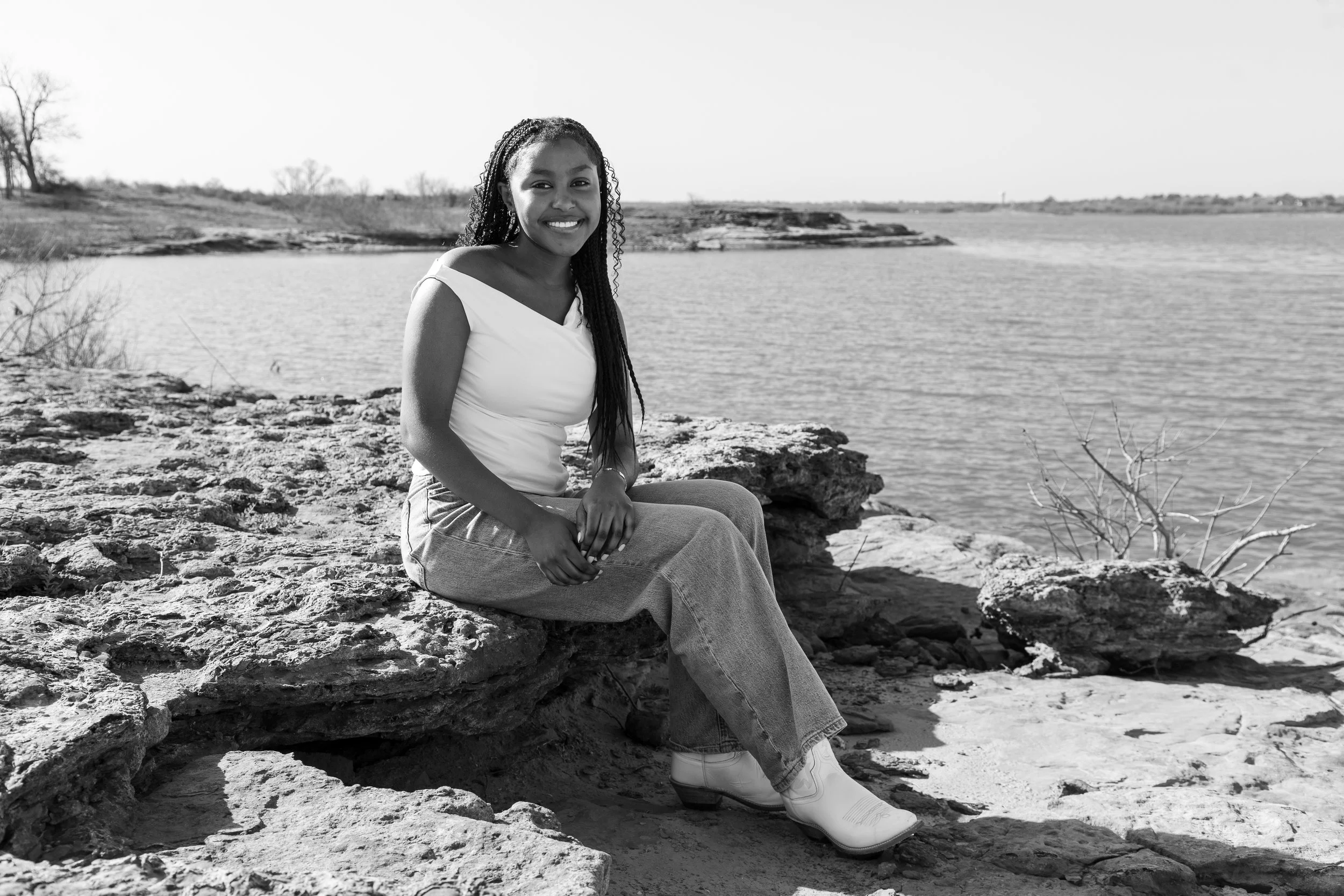 A smiling woman sitting on rocks near a body of water, with a landscape of trees and distant shoreline in the background, in black and white.