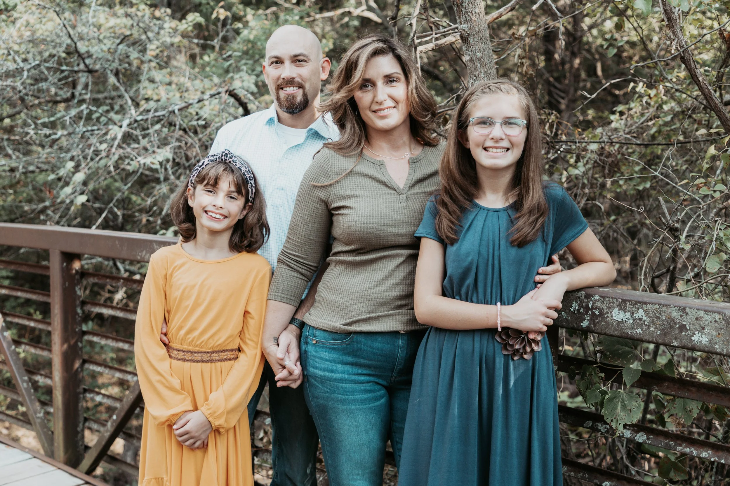 A family of five standing on a wooden bridge outdoors surrounded by trees and foliage. The family includes a mother, father, two teenage girls, and a young girl, all smiling at the camera.