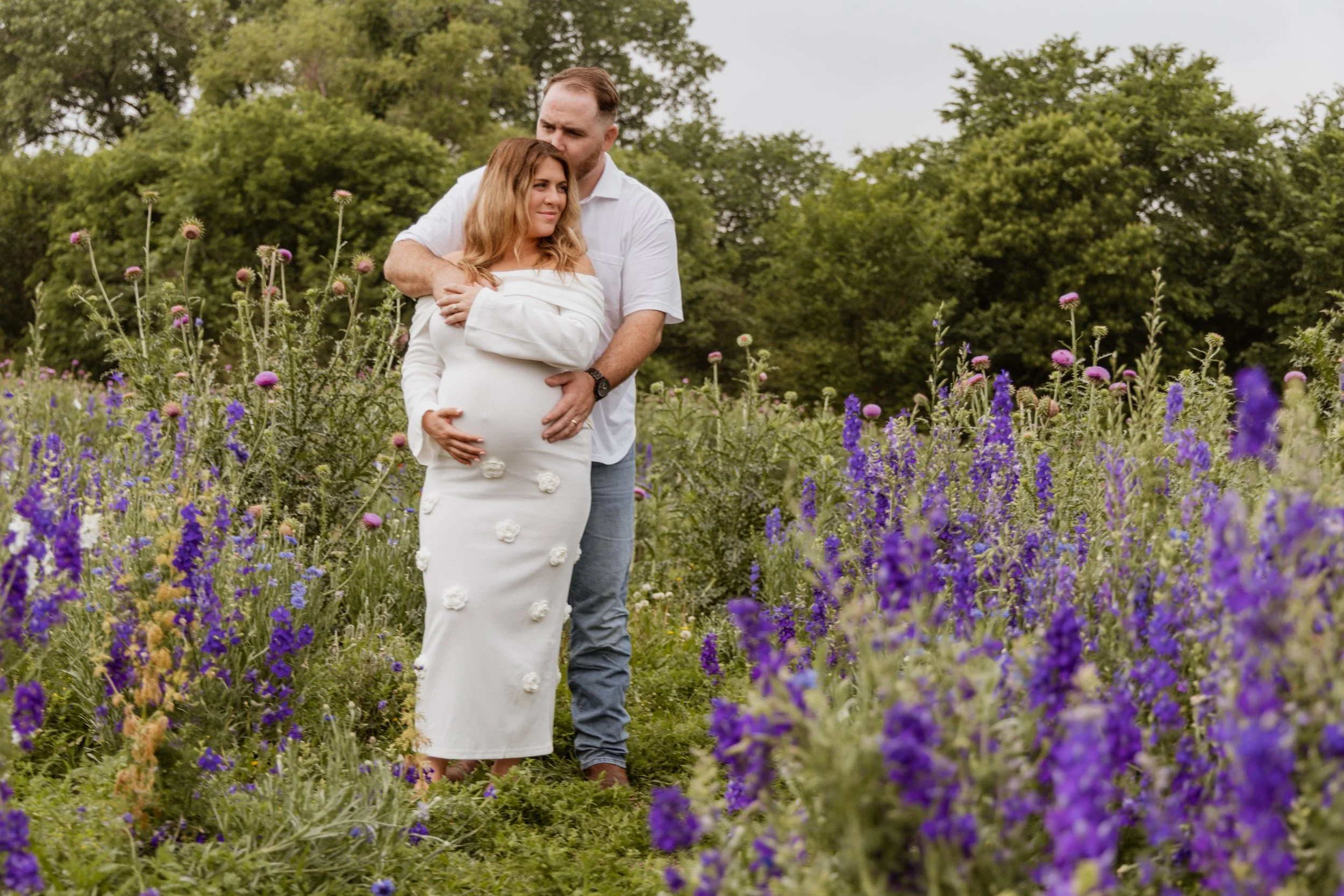 A pregnant woman in a white dress with floral accents standing in a flower field, embracing a man from behind, with trees in the background.