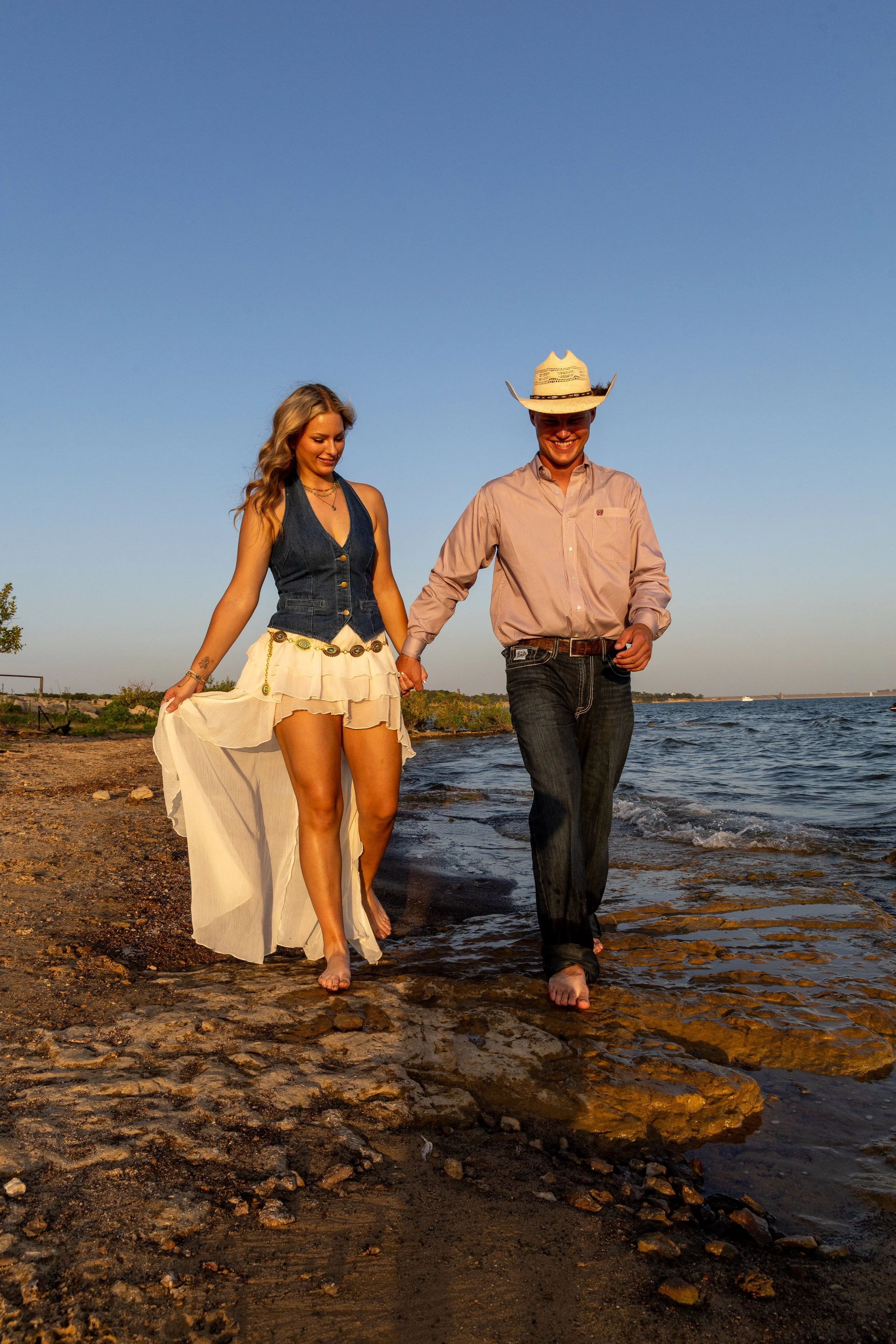 A young woman and man walking barefoot along the shore of a body of water during sunset. The woman wears a denim vest and a flowing skirt, while the man wears a cowboy hat, a long-sleeve shirt, and jeans.