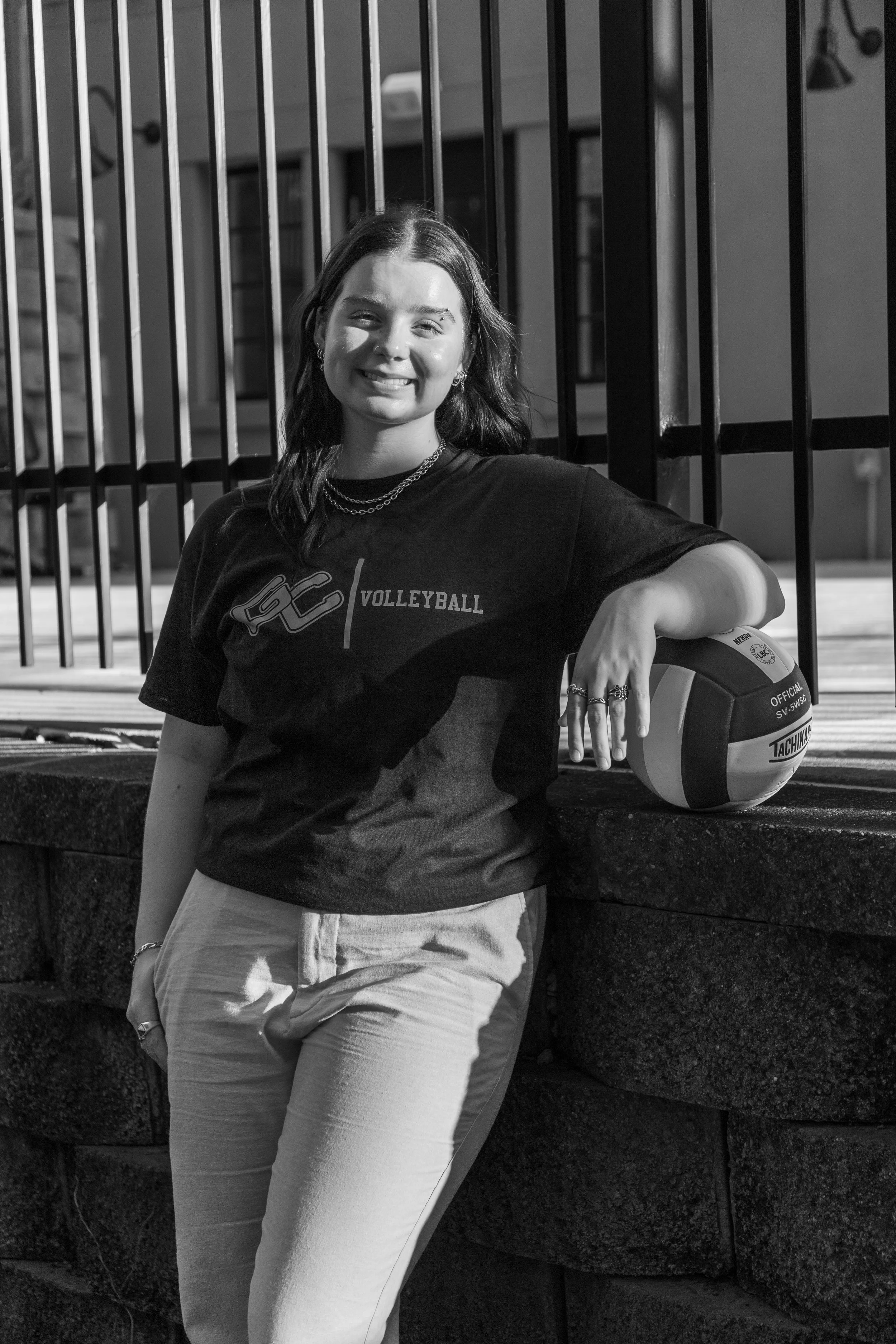 A young woman in volleyball attire smiling and leaning on a volleyball next to a fence.