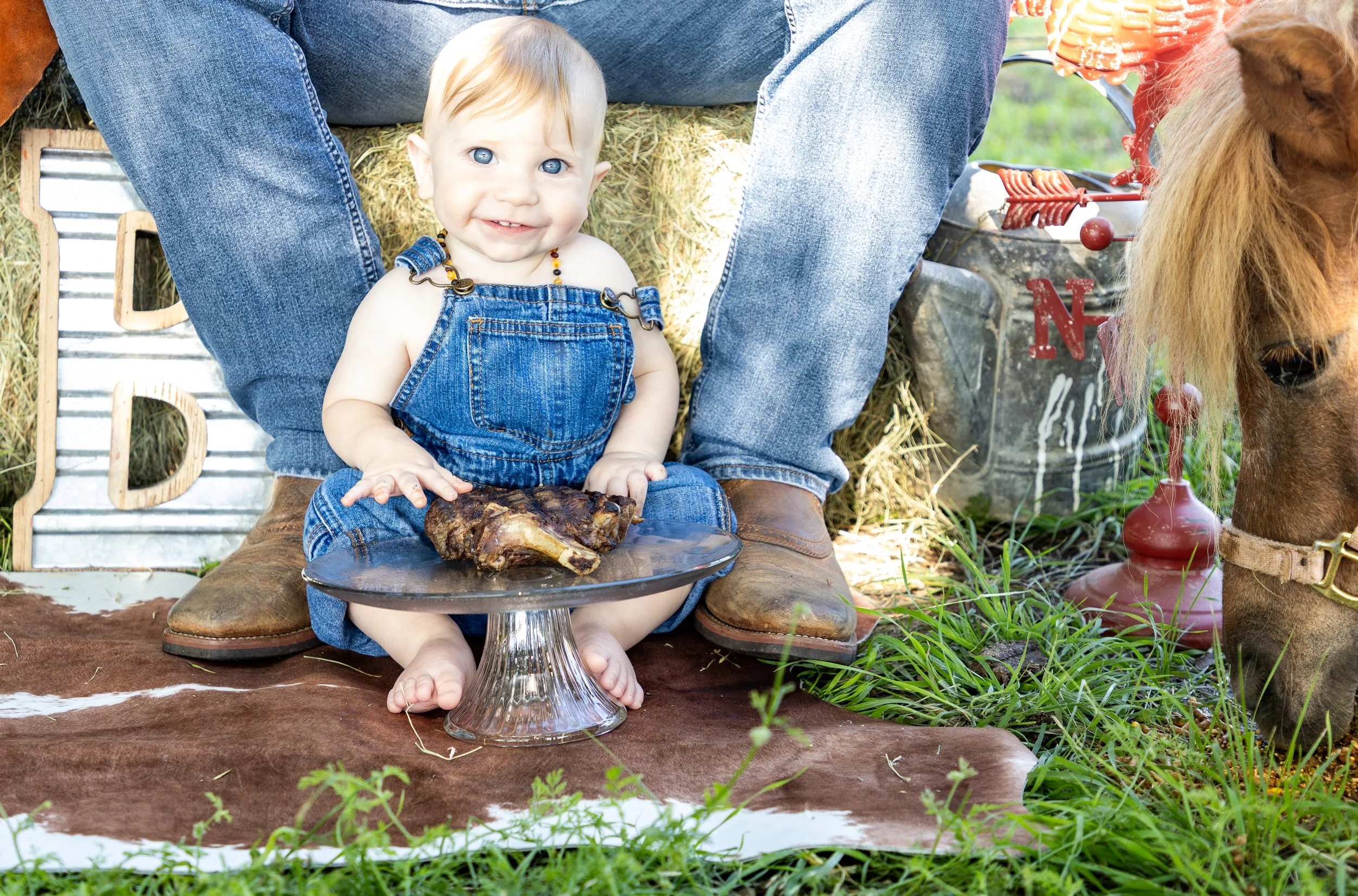 A young child with blue eyes smiling while sitting on a brown and white animal hide, holding a plate with a large piece of cooked meat. The child is dressed in denim overalls. A person wearing jeans and brown boots is sitting behind them, and there's