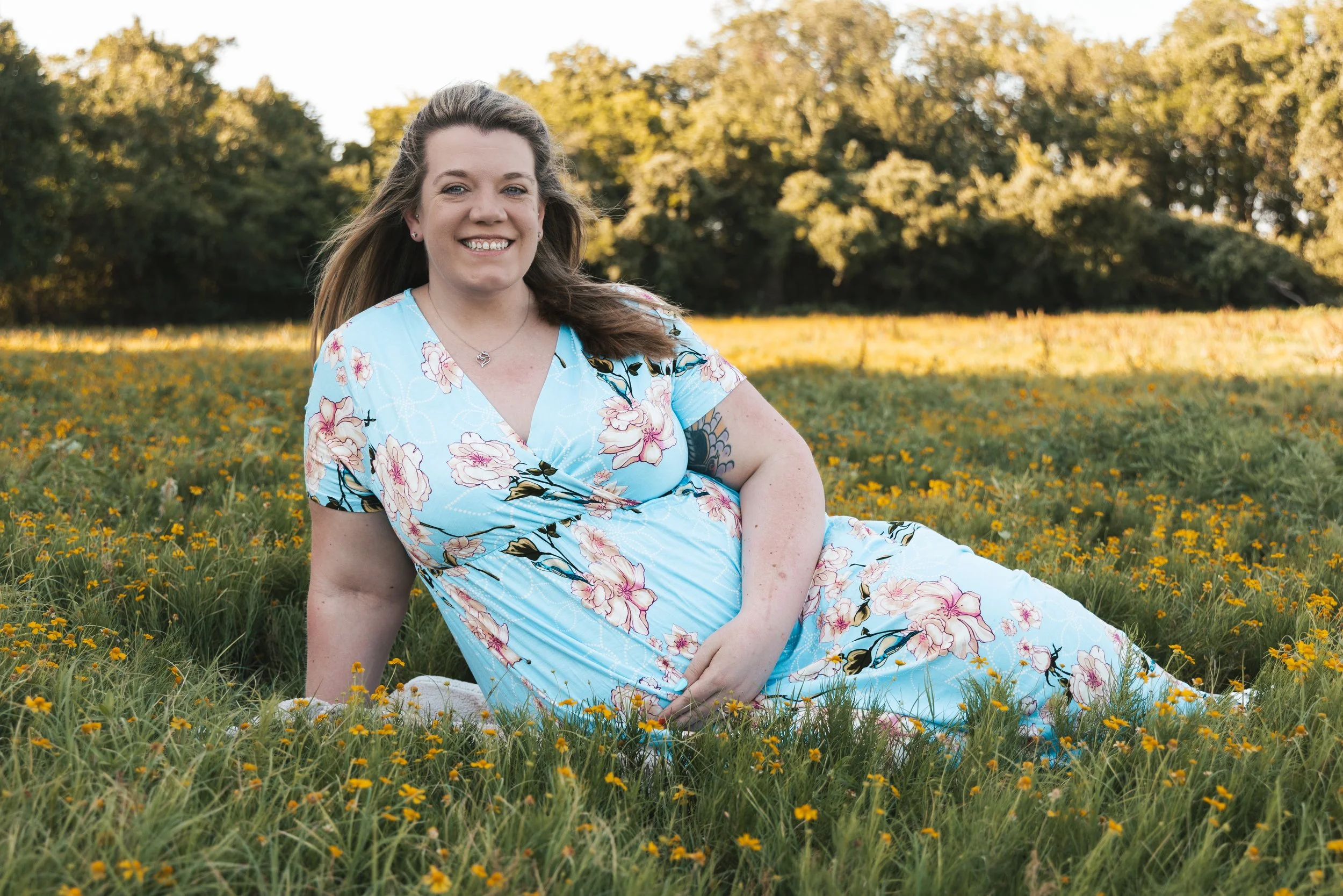 A woman in a floral dress sitting on a field of yellow flowers with trees in the background on a sunny day.