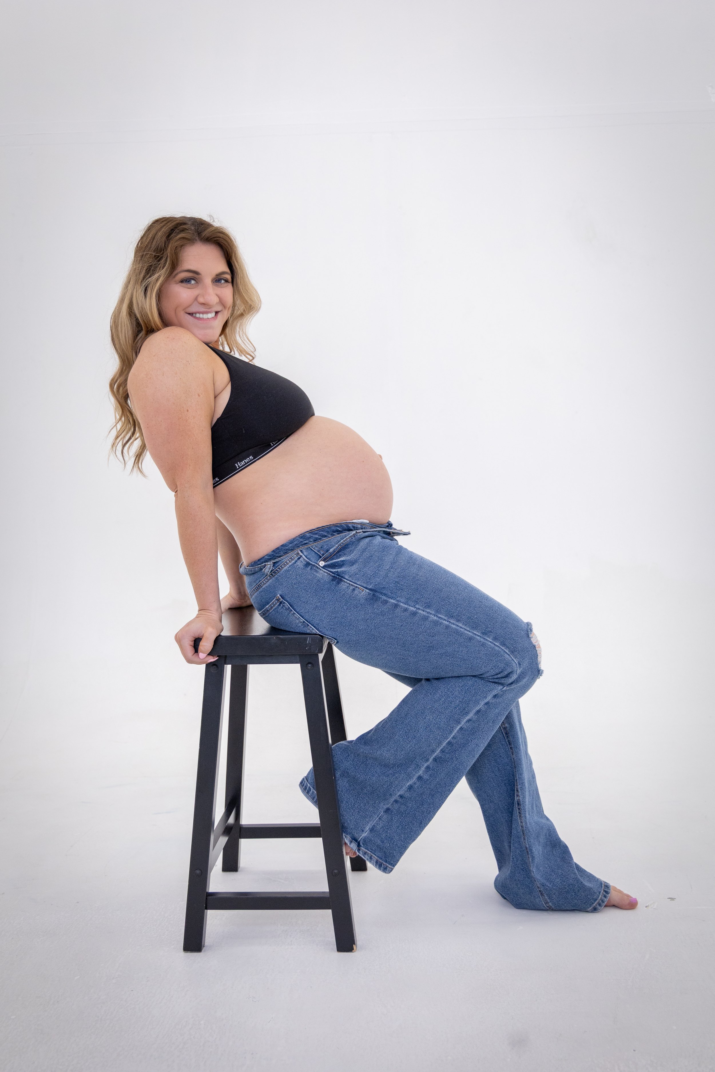 A pregnant woman with long wavy hair, sitting sideways on a black stool, smiling at the camera. She is wearing a black sports bra and blue jeans with a rip on one knee, against a plain white background.