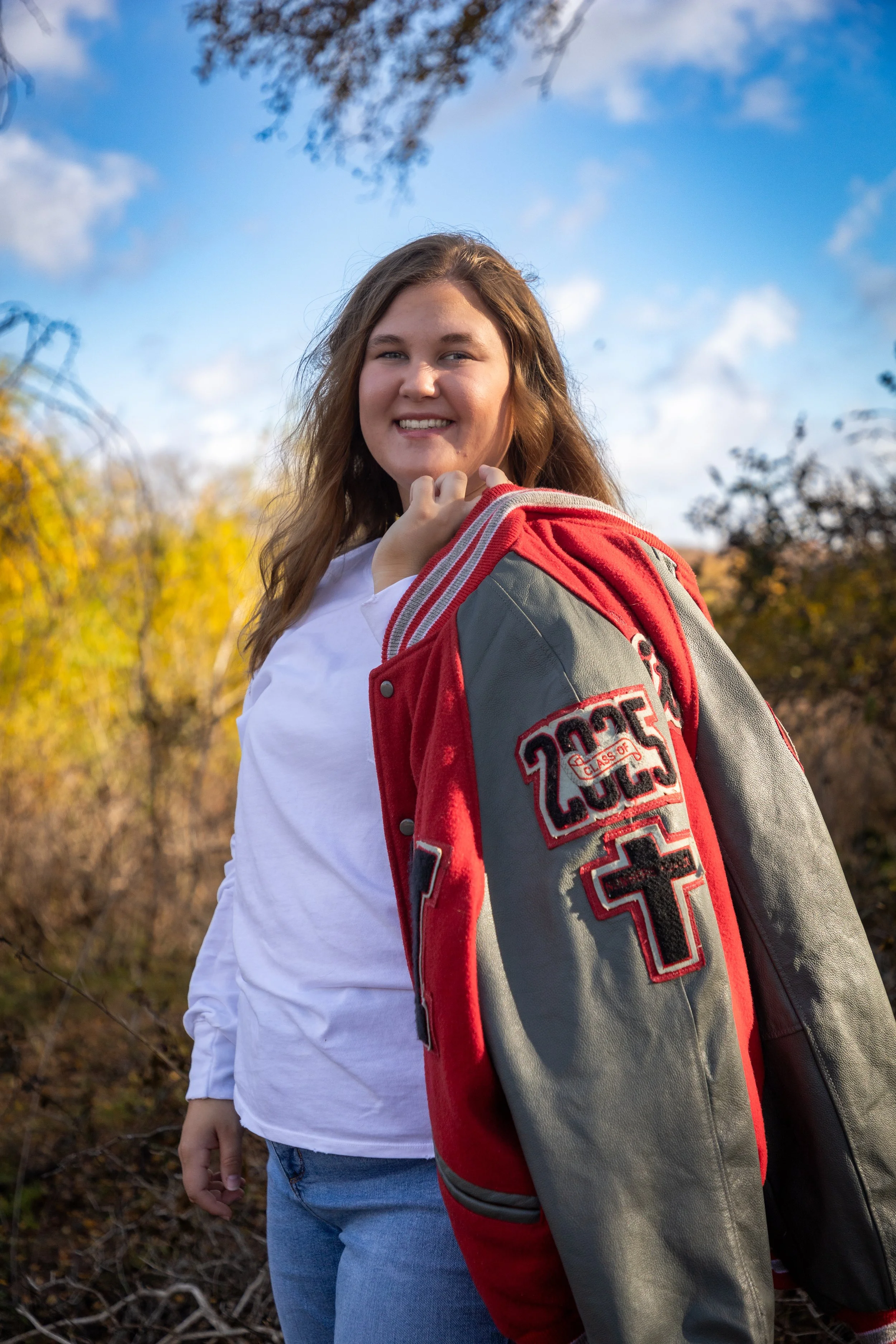 Young woman outdoors with a smile, wearing a red and gray varsity jacket over a white shirt, standing in front of autumn trees with blue sky in the background.