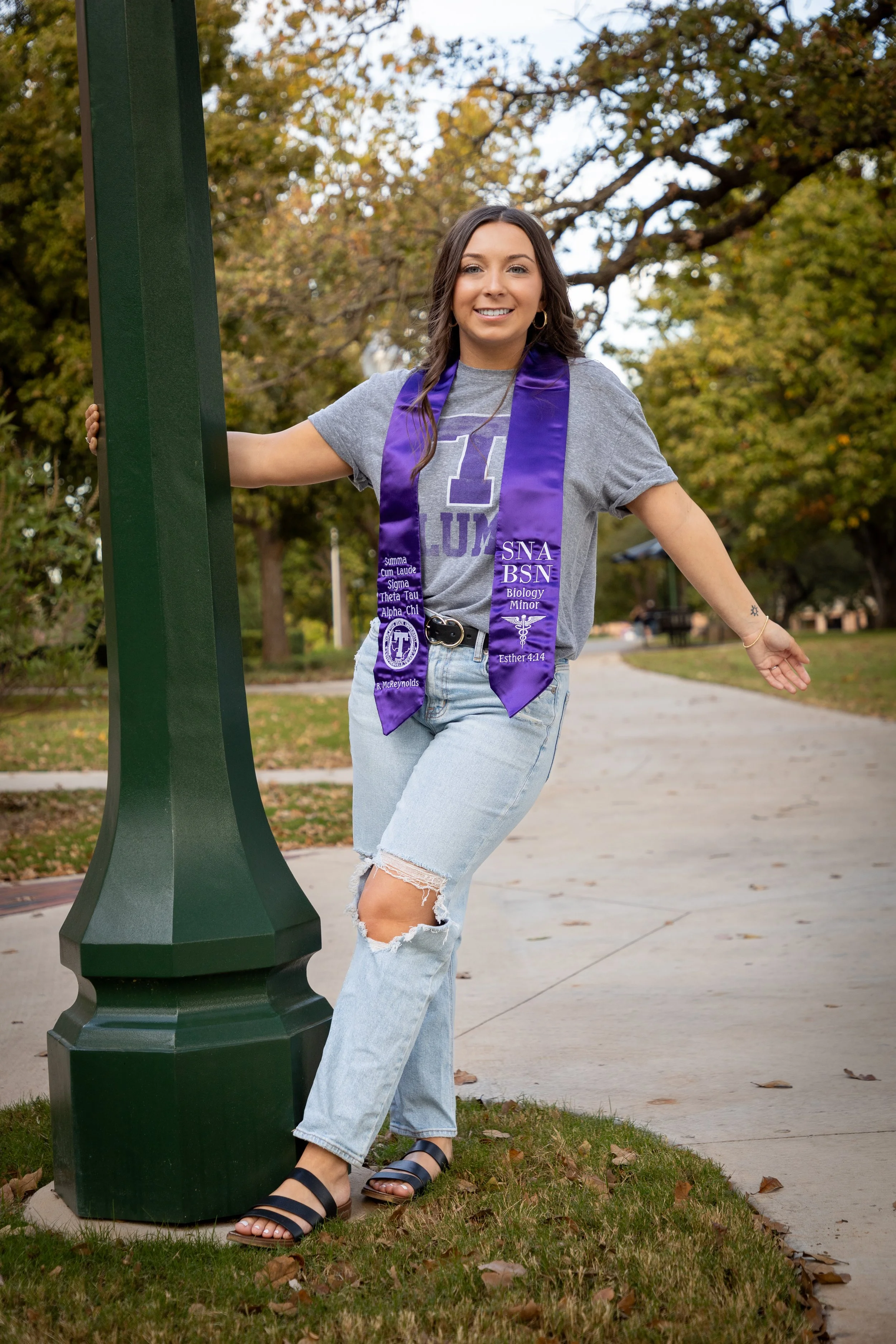 A young woman wearing a gray T-shirt with Greek letters and a purple graduation stole, standing outdoors on a sidewalk, posing with one arm on a green lamppost and smiling at the camera. She has long brown hair, ripped jeans, and sandals, with fall t
