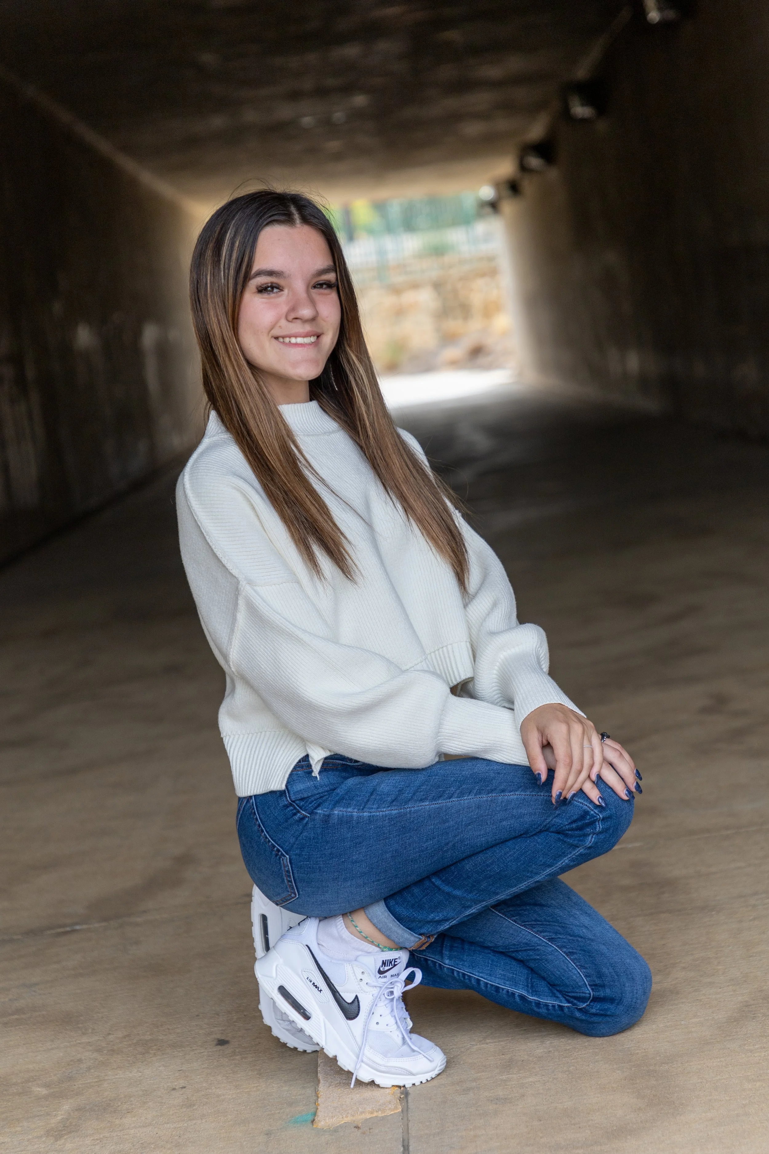 A young woman with long brown hair, wearing a white sweater, blue jeans, and white Nike sneakers, kneeling on the ground in an underpass, smiling at the camera.