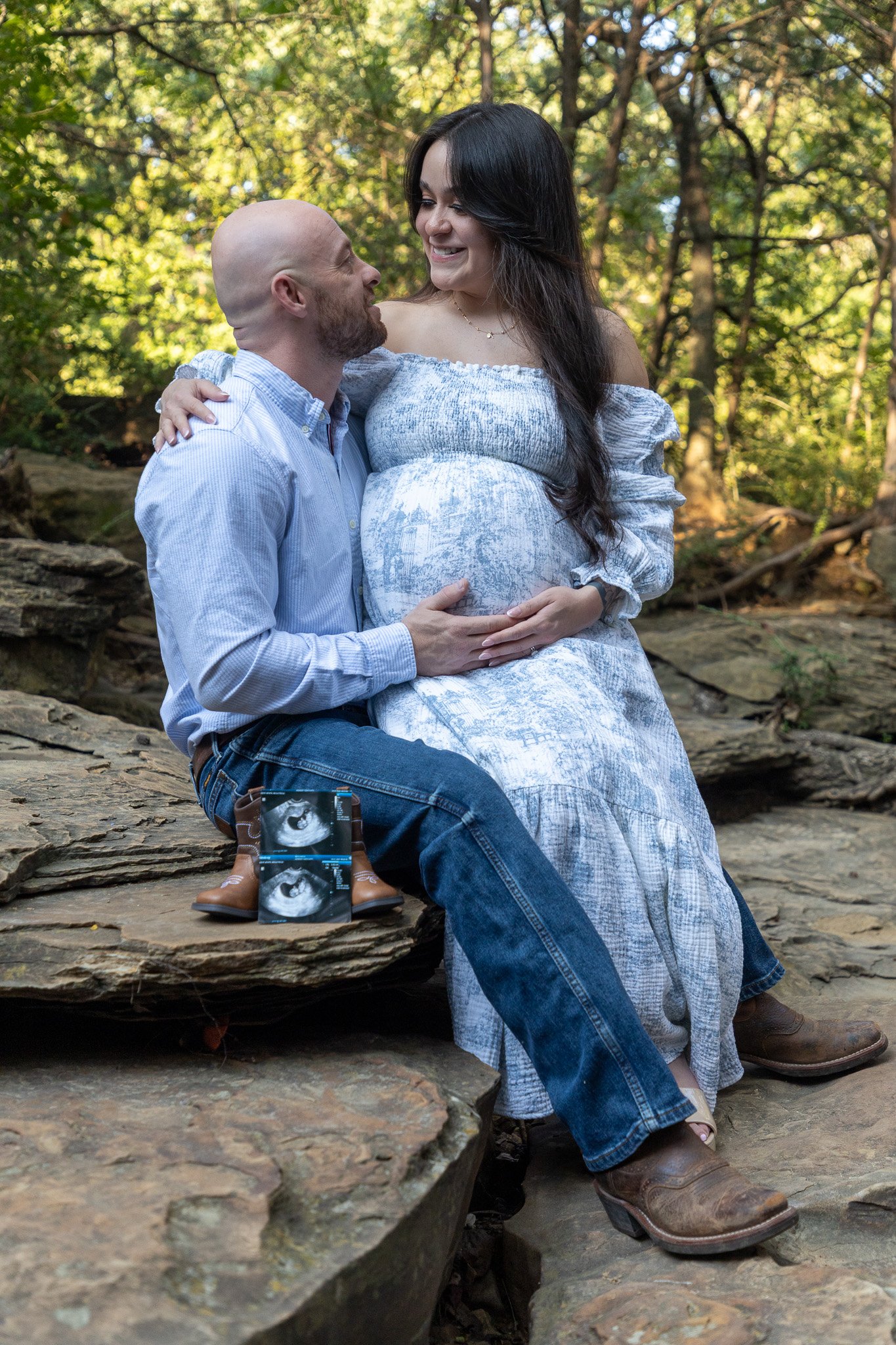 A couple sitting on rocks outdoors, with the woman pregnant, smiling at each other. There are ultrasound images placed on her lap. The background features trees and greenery.