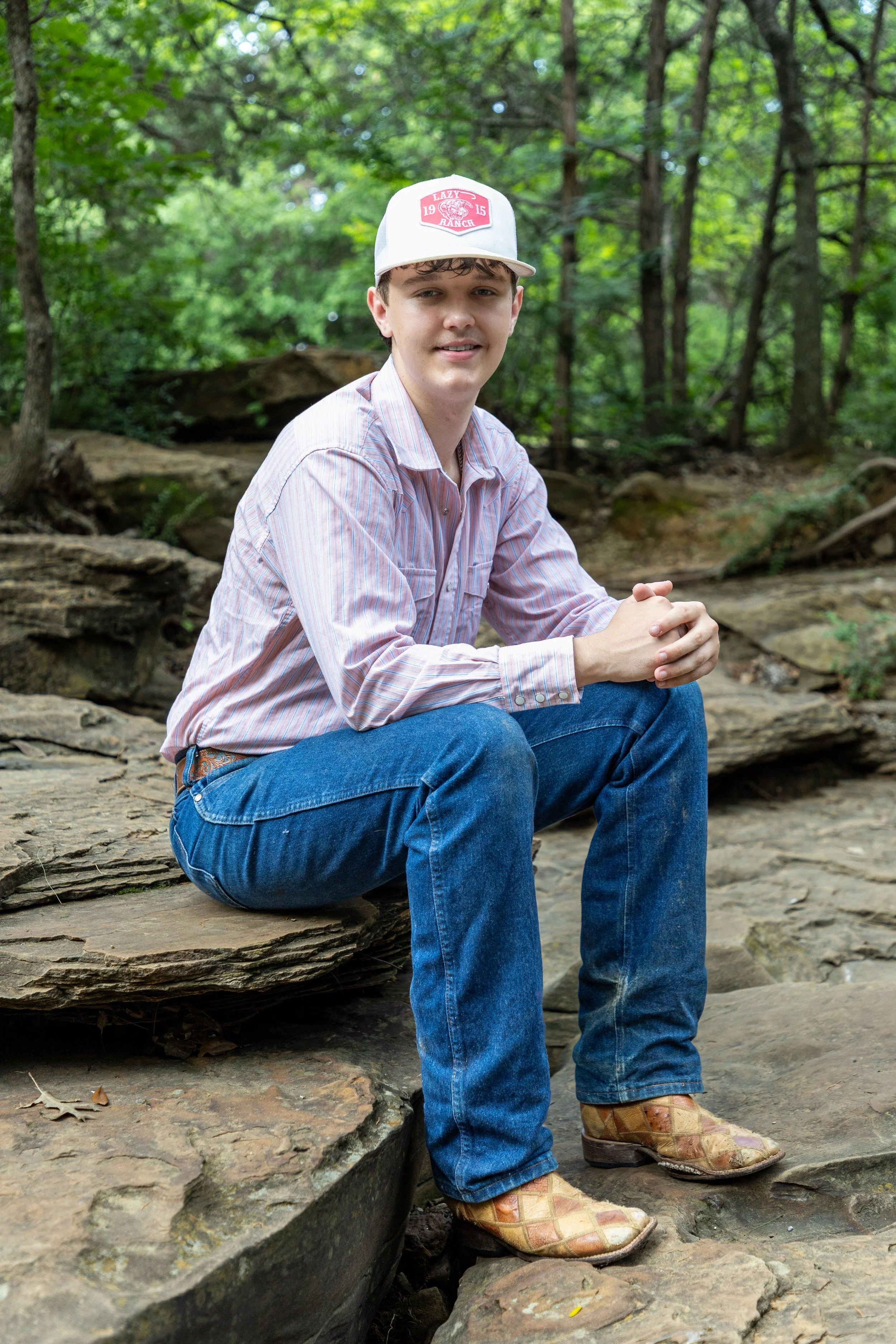 A young man sitting on rocks in a forest, wearing a white cap, pink striped shirt, jeans, and tan cowboy boots.