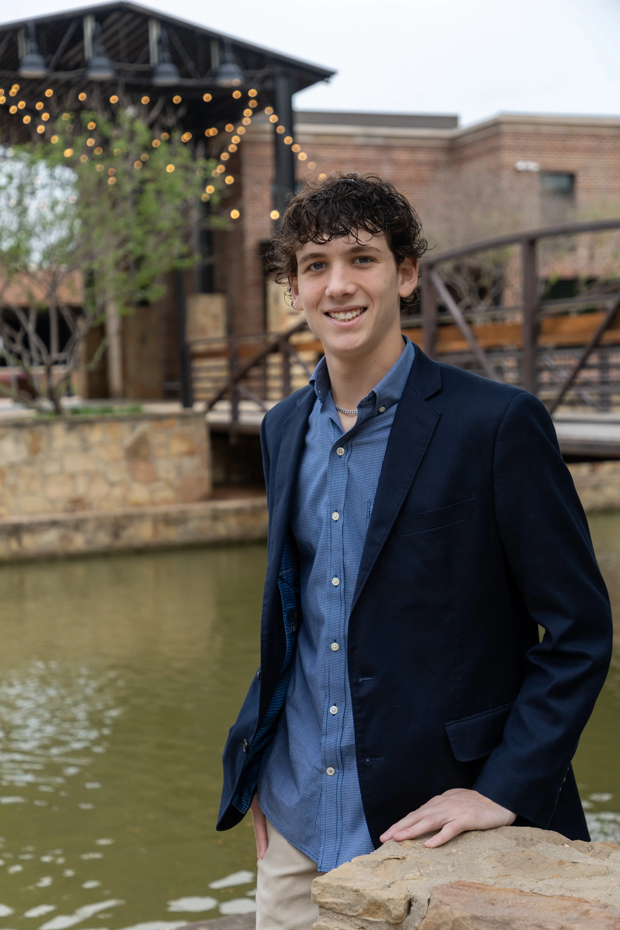 A young man with curly brown hair in a navy blazer and blue shirt, smiling, standing by a small river with a stone wall and a wooden bridge in the background, decorated with string lights.