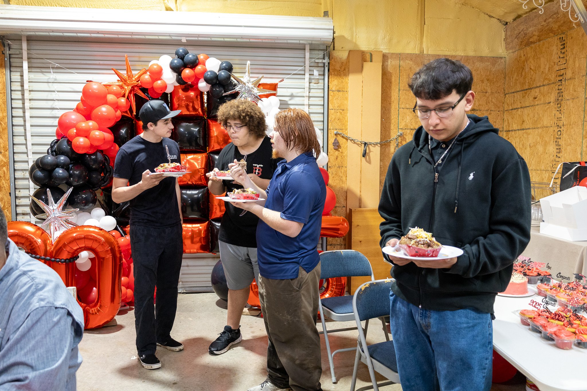 People at a celebration, standing near a balloon decoration with black, red, and white balloons, and a table with cupcakes.