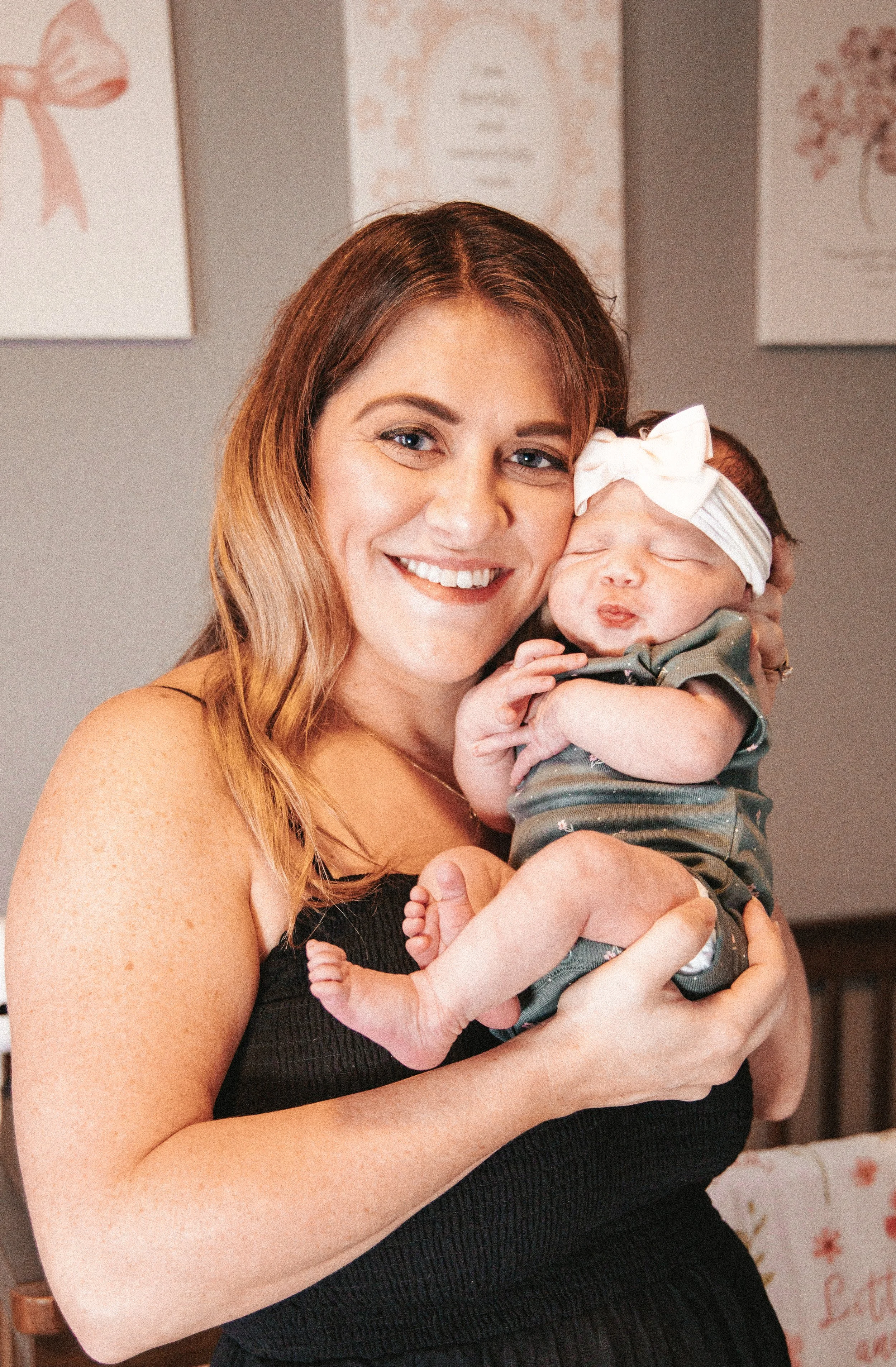 A woman with long, wavy, auburn hair smiles while holding a sleeping newborn baby, who is wearing a large white bow headband and a dark, patterned outfit. The background features decorated walls with pink floral and ribbon designs.
