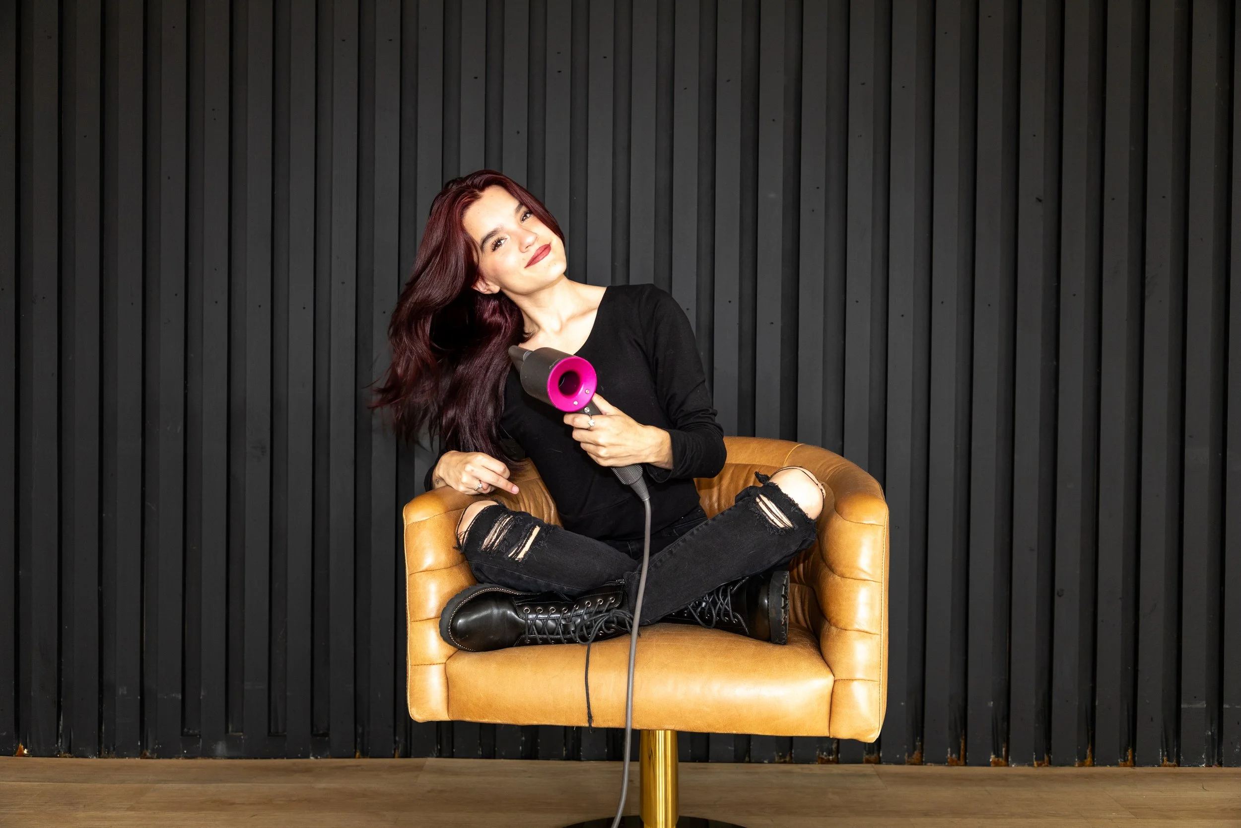 Woman with red hair sitting cross-legged on a tan armchair, using a hair dryer, against a black striped wall background.