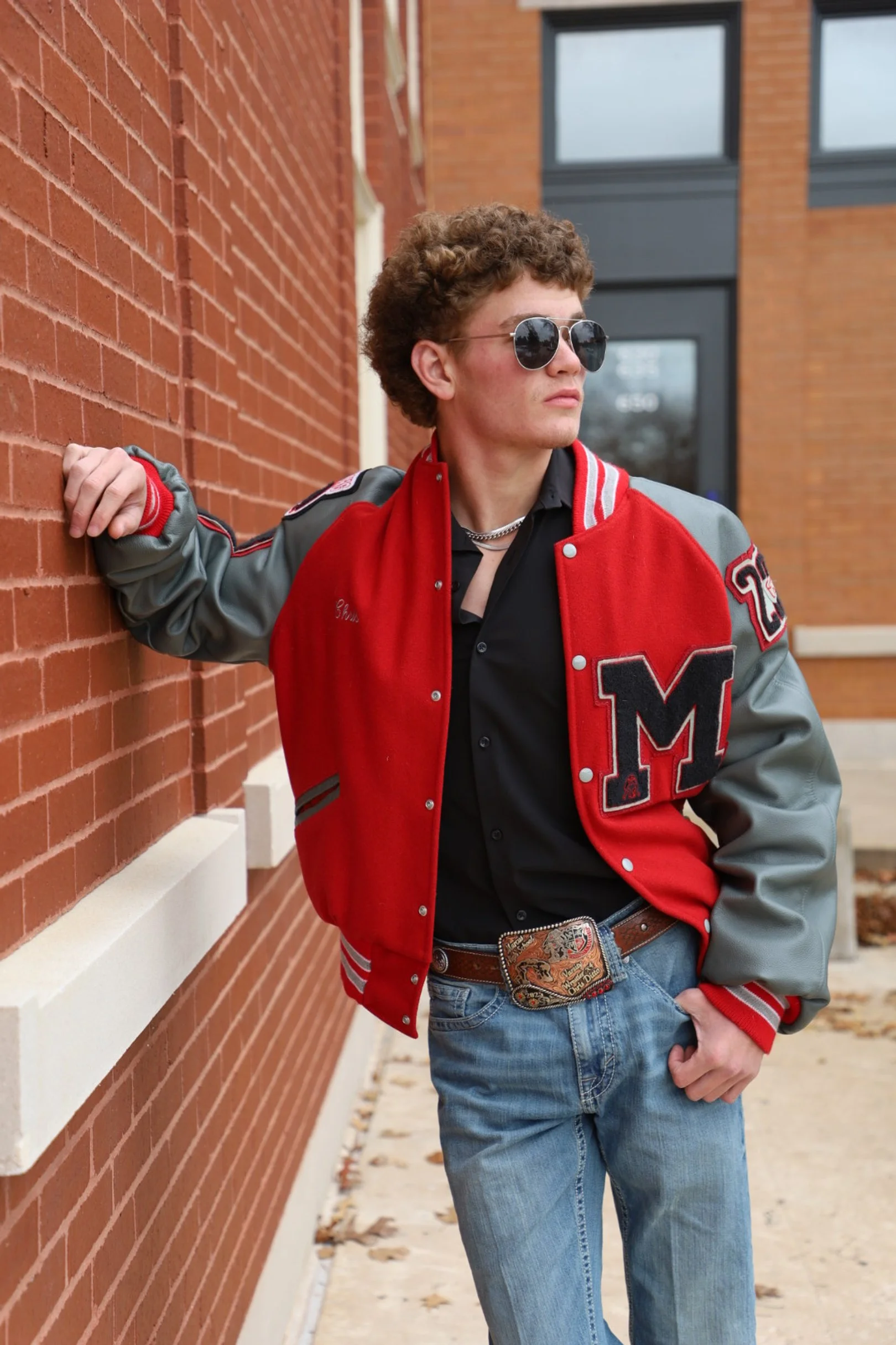 Young man with curly hair wearing sunglasses, a red and gray varsity jacket, black shirt, blue jeans, standing against a brick wall.