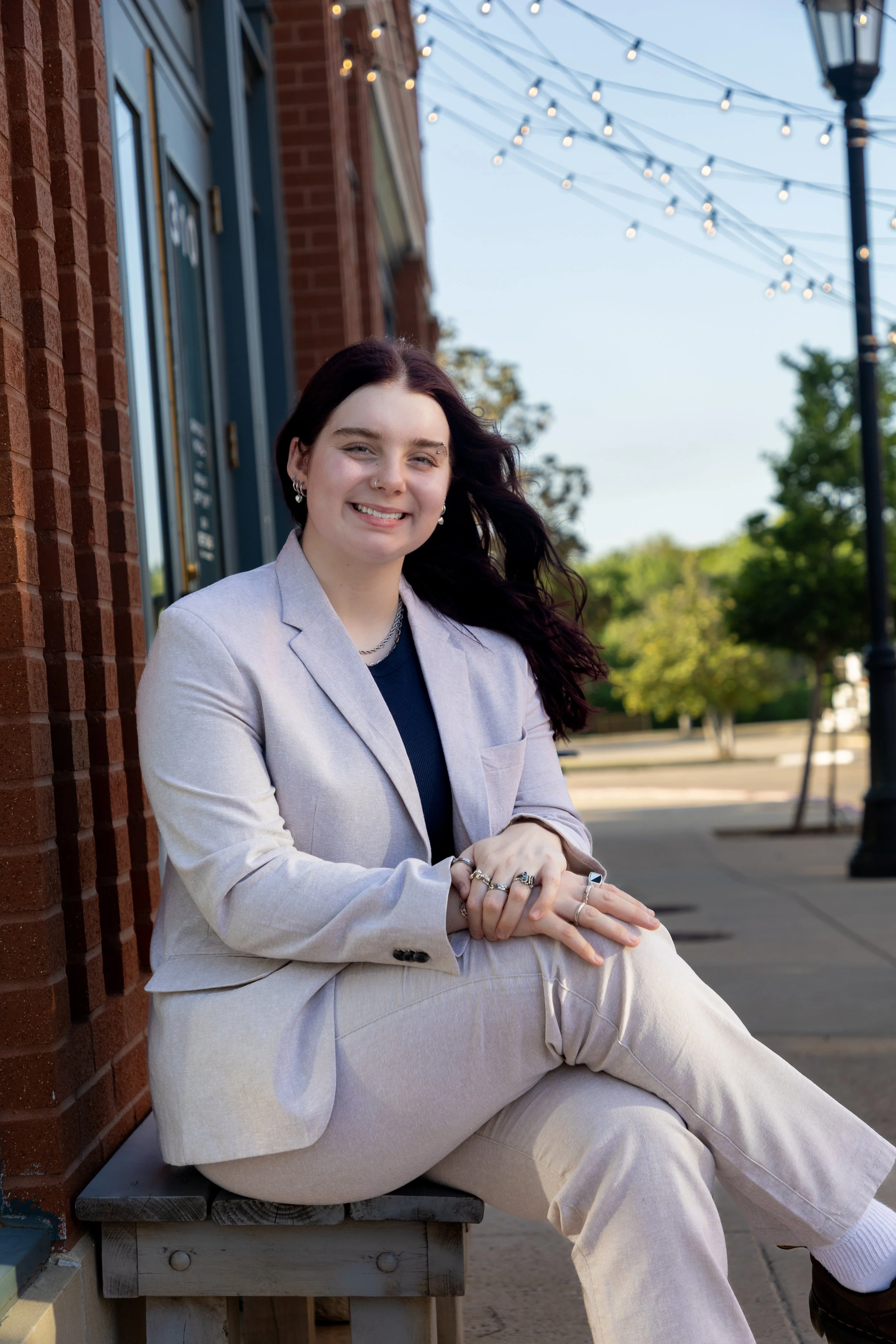 A woman with dark hair wearing a light-colored suit with a black shirt sitting on a bench outside on a sidewalk, smiling at the camera, with string lights and trees in the background.