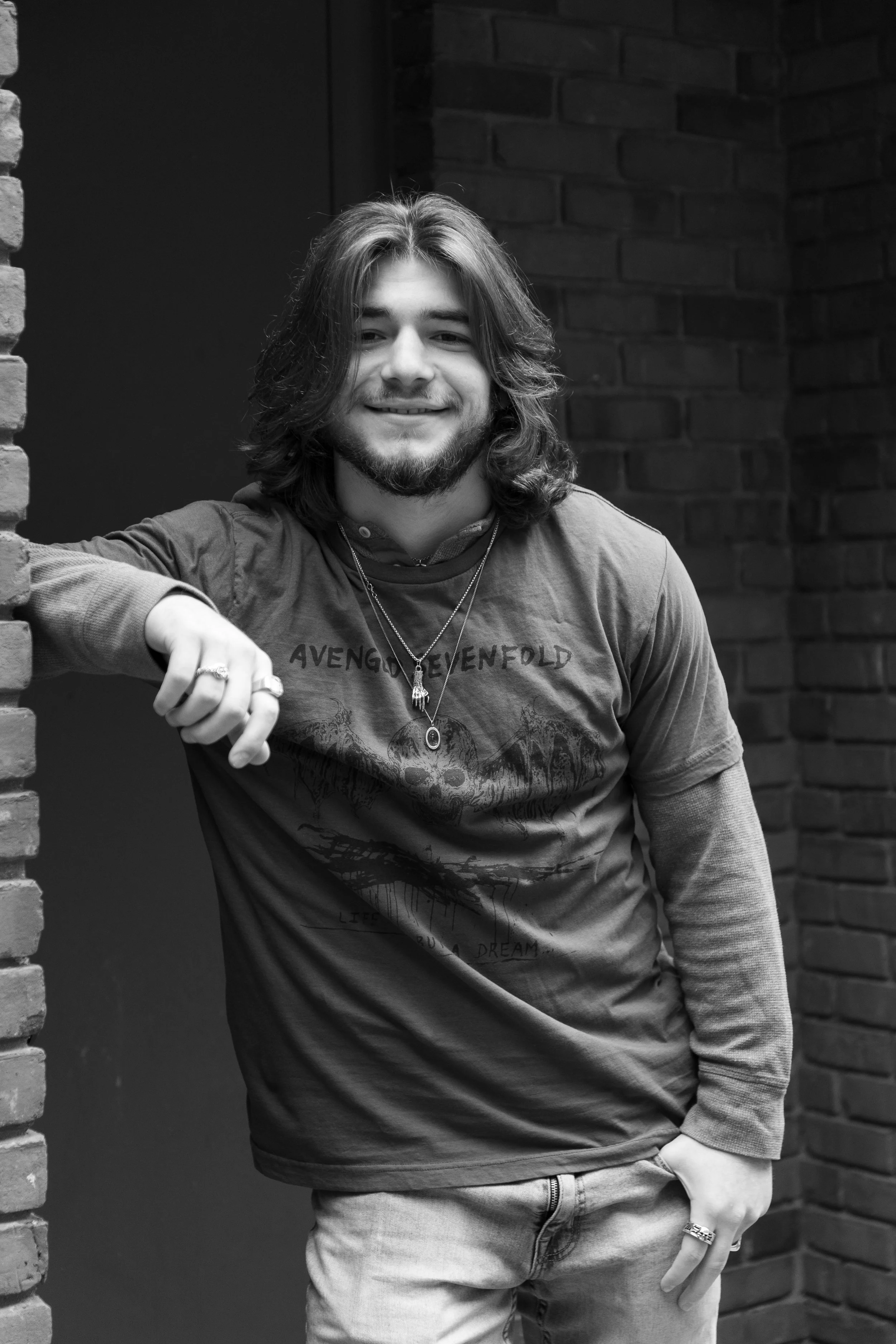A young man with long hair, a beard, and a smile, leaning against a brick wall in black and white.