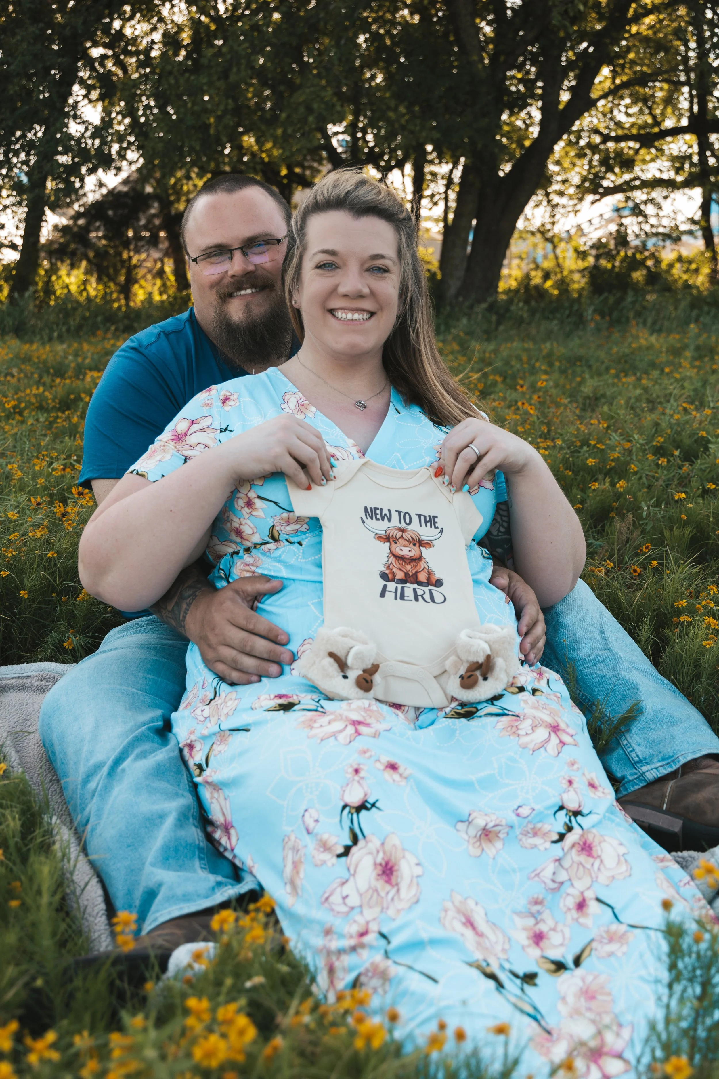 A smiling woman sitting on a blanket holding a baby onesie with a cartoon animal and the text "New to the Herd", with a man behind her in a grassy field with yellow flowers and trees in the background.