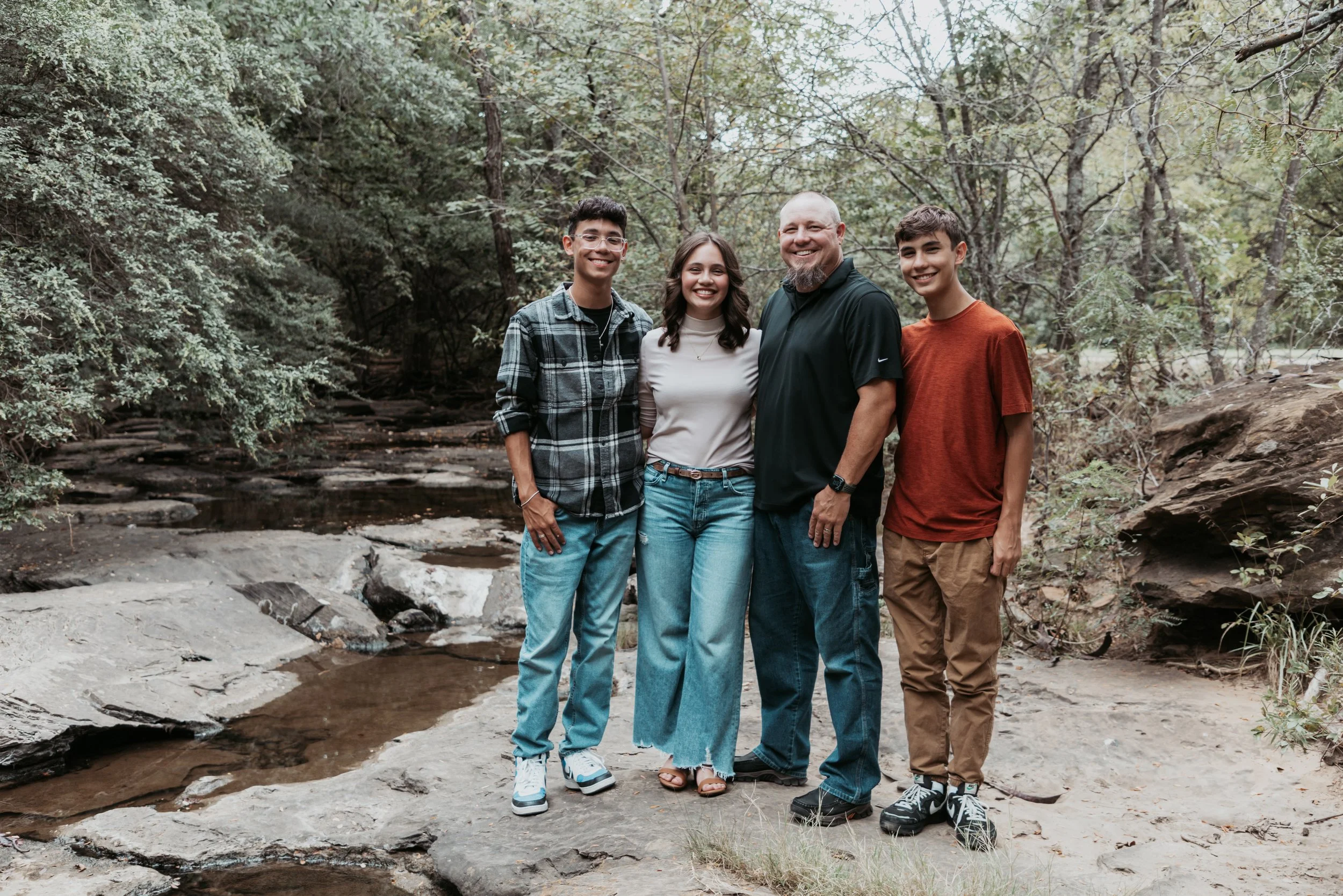 Group of five people standing outdoors near a stream in a forest, smiling for the camera.