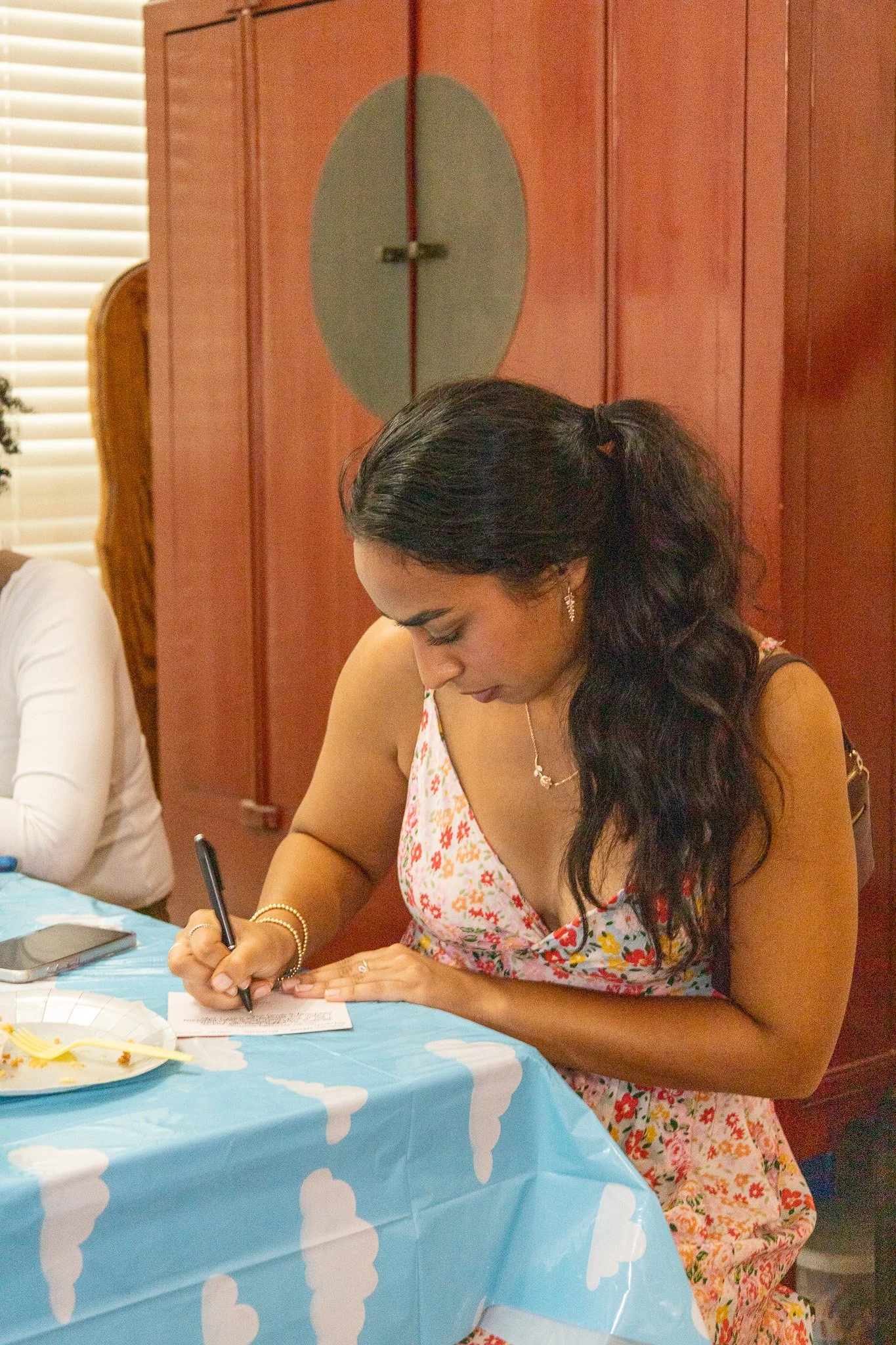 A woman in a floral dress with long dark hair tied back writes on a piece of paper at a table with a blue cloud-patterned tablecloth during a celebration.