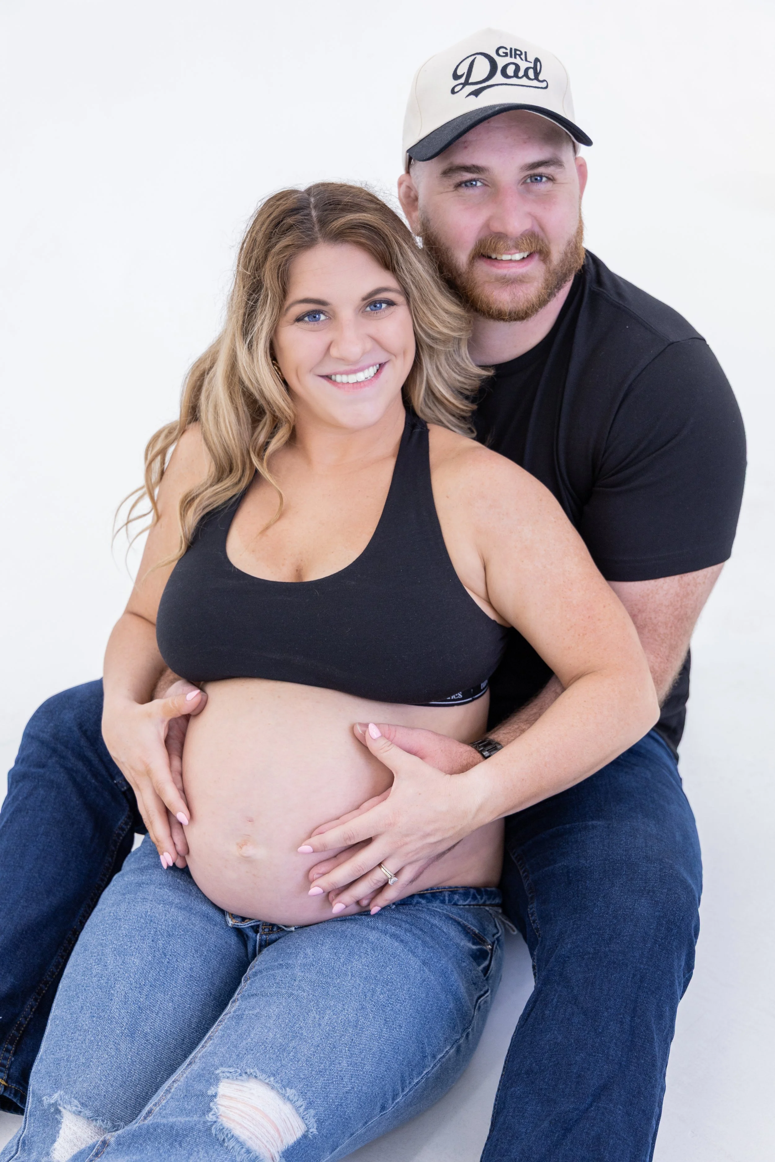 A happy pregnant woman with long wavy hair, wearing a black sports bra and ripped jeans, sitting on her partner's lap and holding her belly. Her partner, a bearded man with blue eyes, wearing a black t-shirt, blue jeans, and a white cap that says 'Gi