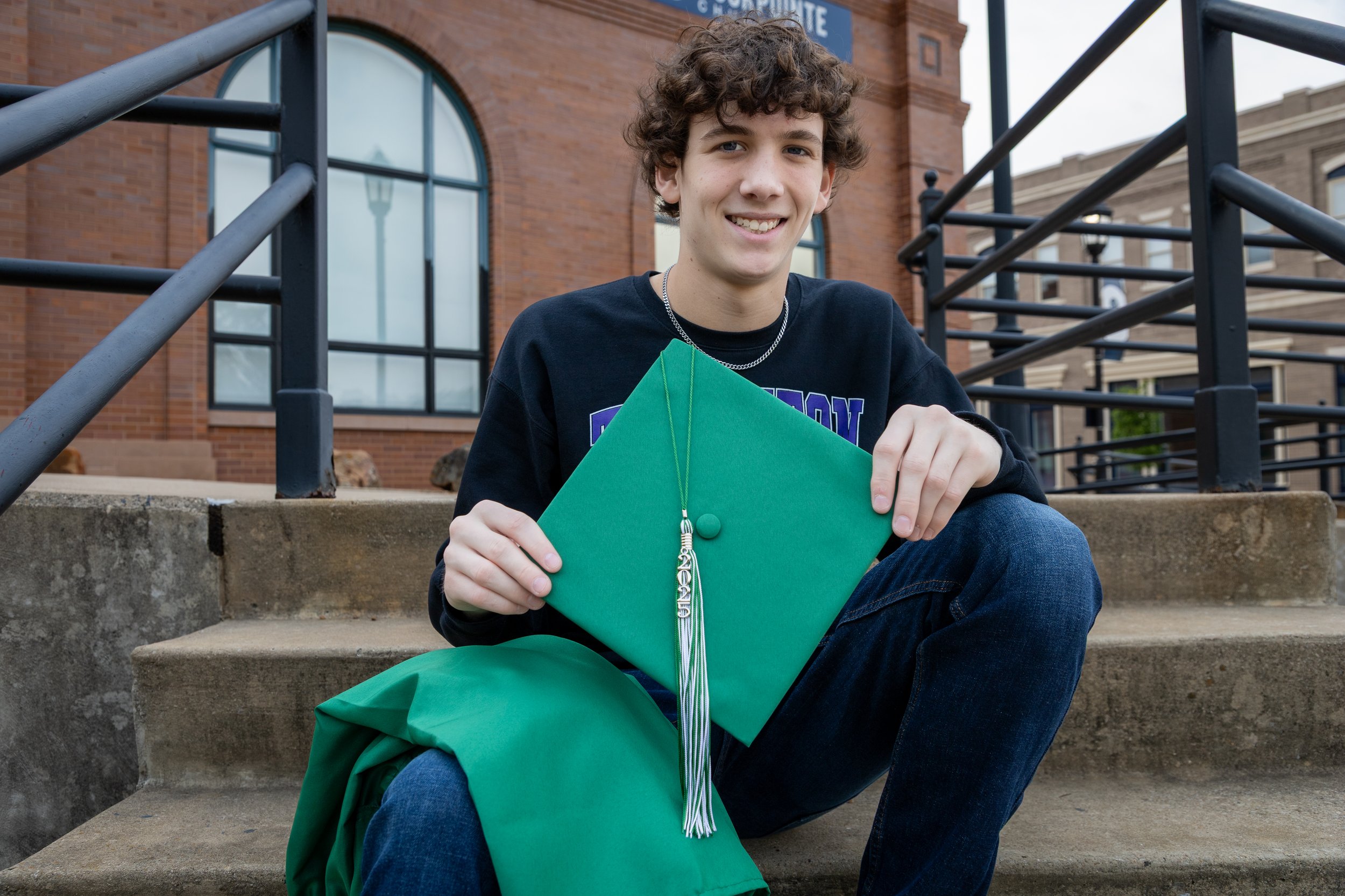 Young man sitting on the steps in front of a brick building, holding a green graduation cap and gown, smiling at the camera.