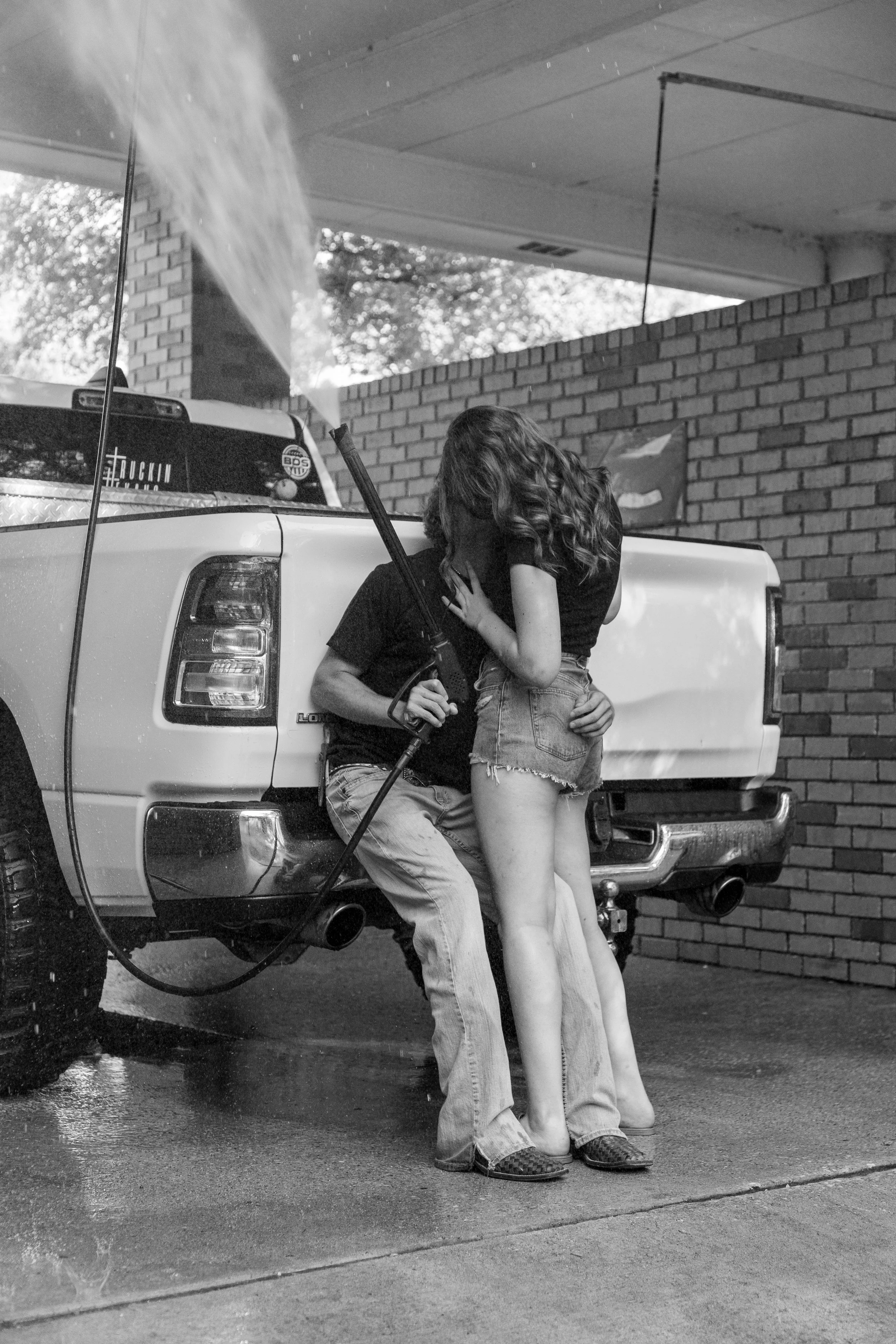 A black and white photo of a man and woman kissing at a car wash, the man sitting on the back of a pickup truck holding a garden hose, woman standing on the ground with her arm around his neck.