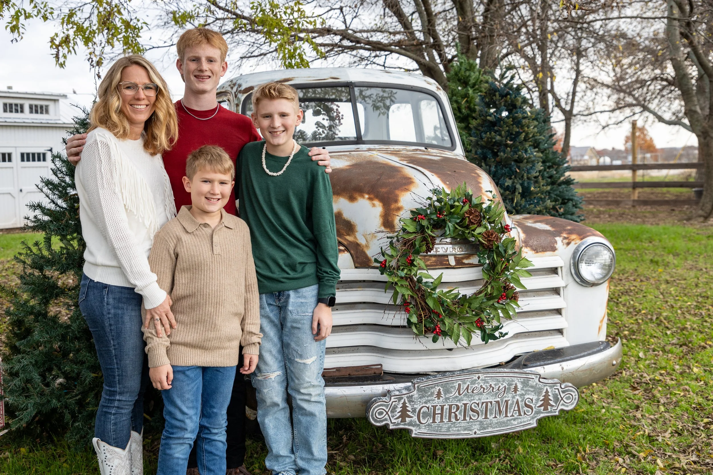 A woman and four boys are standing outdoors in front of a rusty vintage car decorated with a Christmas wreath. There are Christmas trees and a sign that says 'Merry Christmas' behind them.