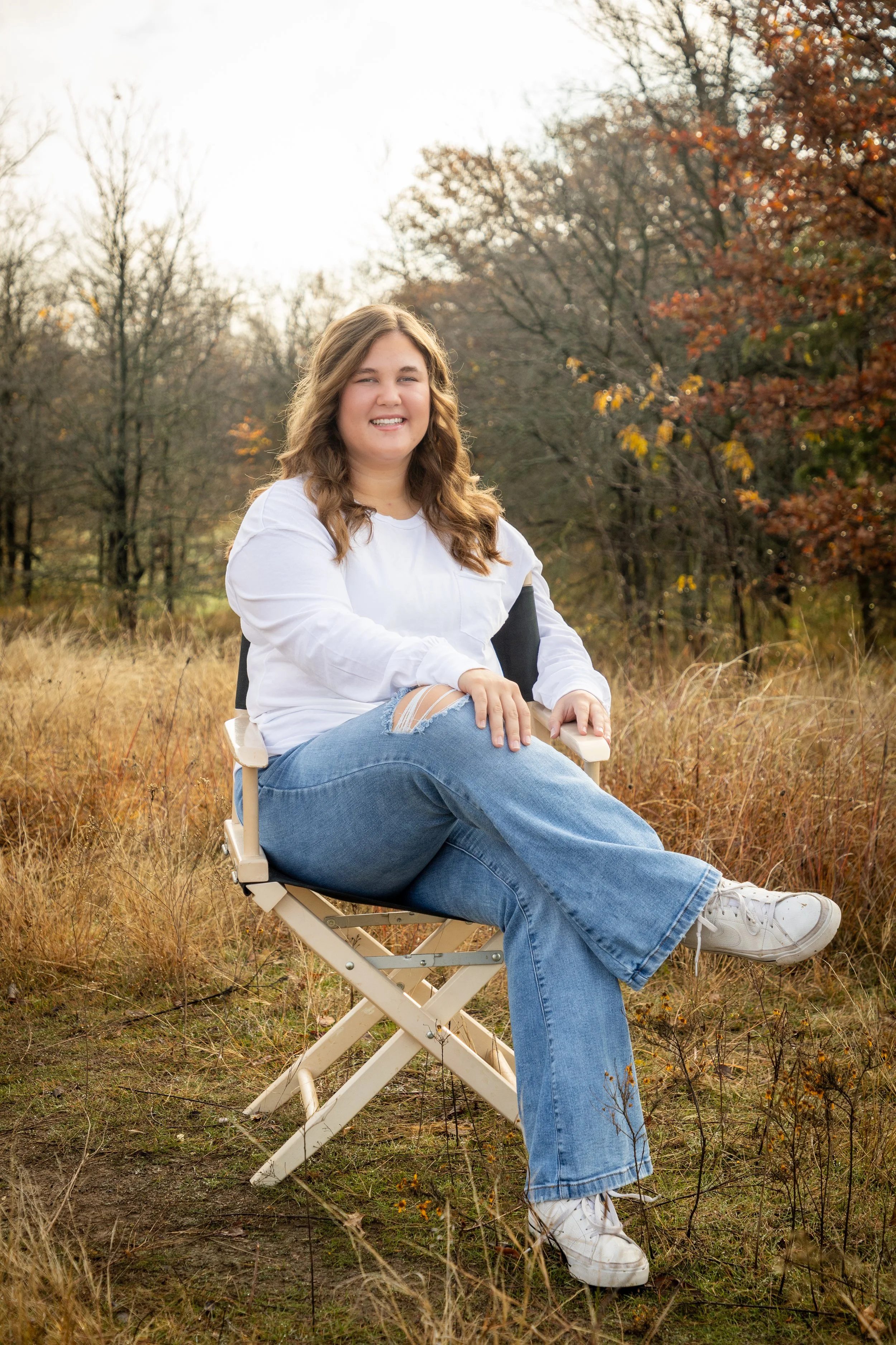 A young woman with light brown hair sitting on a foldable chair in a grassy field during fall, smiling at the camera with trees in the background.
