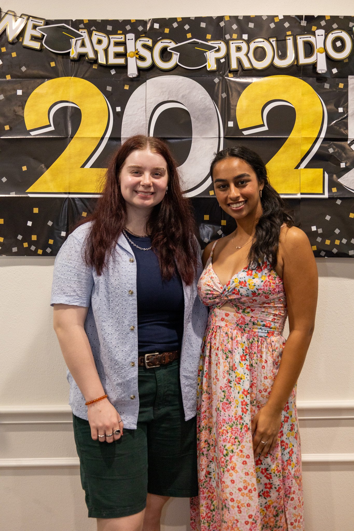 Two young women standing tightly side-by-side, smiling, during a graduation celebration. Behind them is a large black banner with yellow, white, and gray lettering that reads "We Are So Proud of You" and a large "2022" sign.