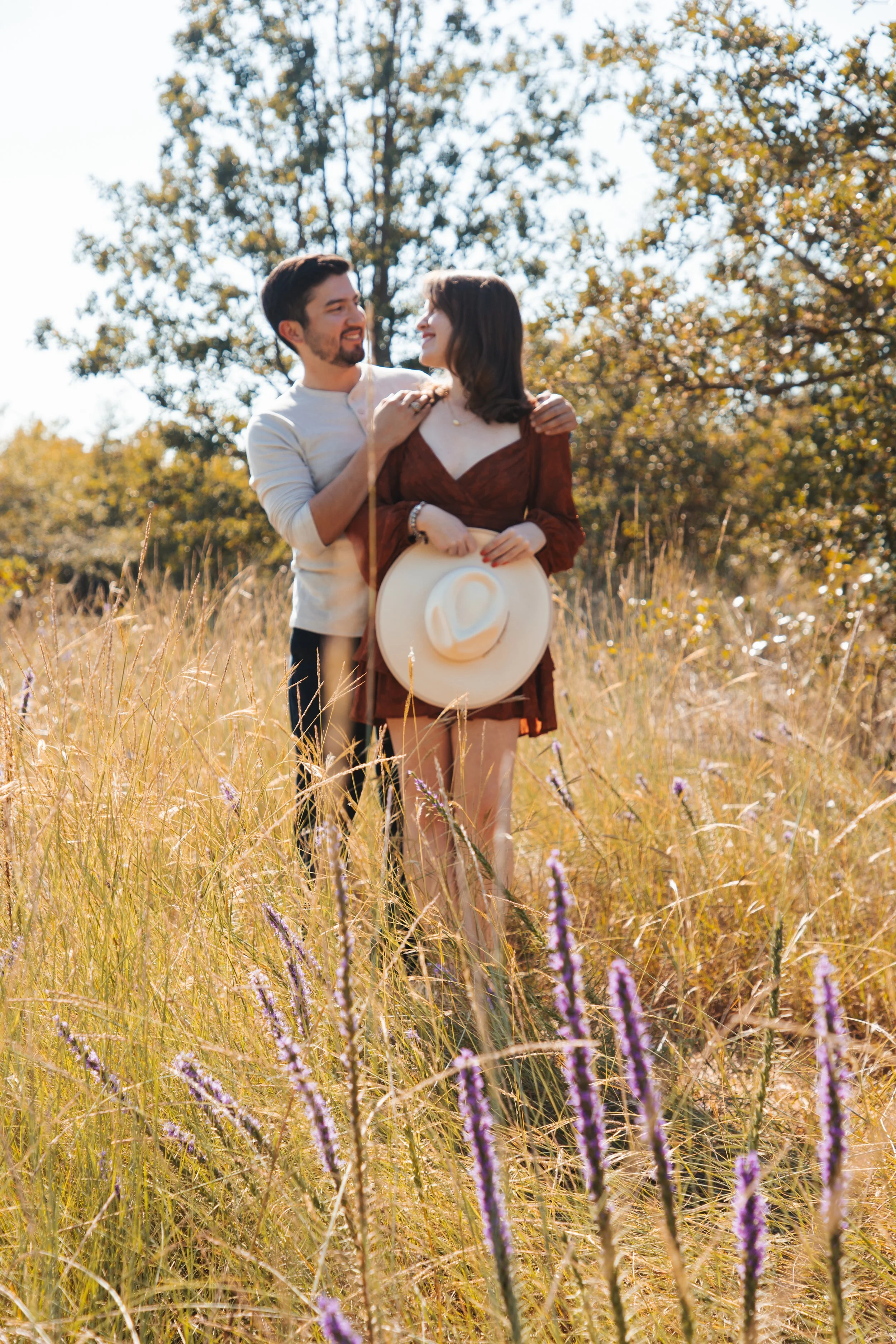 A couple standing in a field of tall grass and purple flowers on a sunny day, smiling and looking at each other. The woman is holding a wide-brimmed hat, wearing a reddish-brown dress, and the man is in a light-colored shirt.
