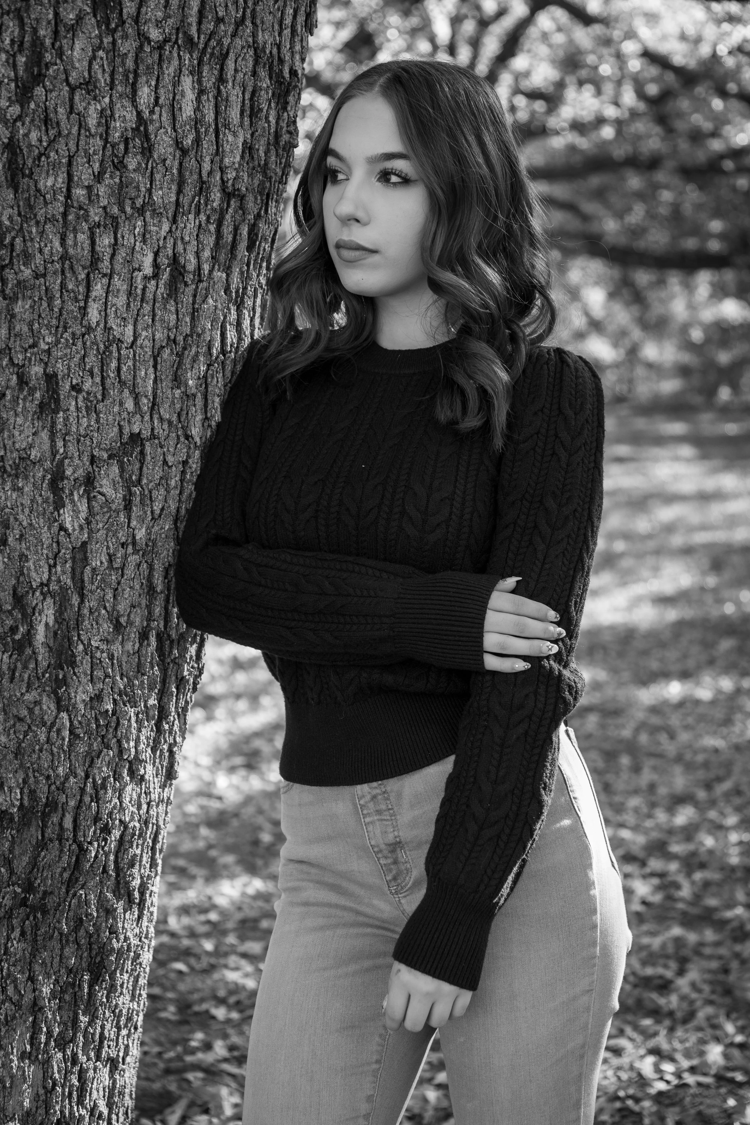 A black and white photo of a young woman with wavy hair standing next to a tree, wearing a dark cable-knit sweater and light-colored jeans, with her arms crossed and gazing thoughtfully to the side in a park-like setting.