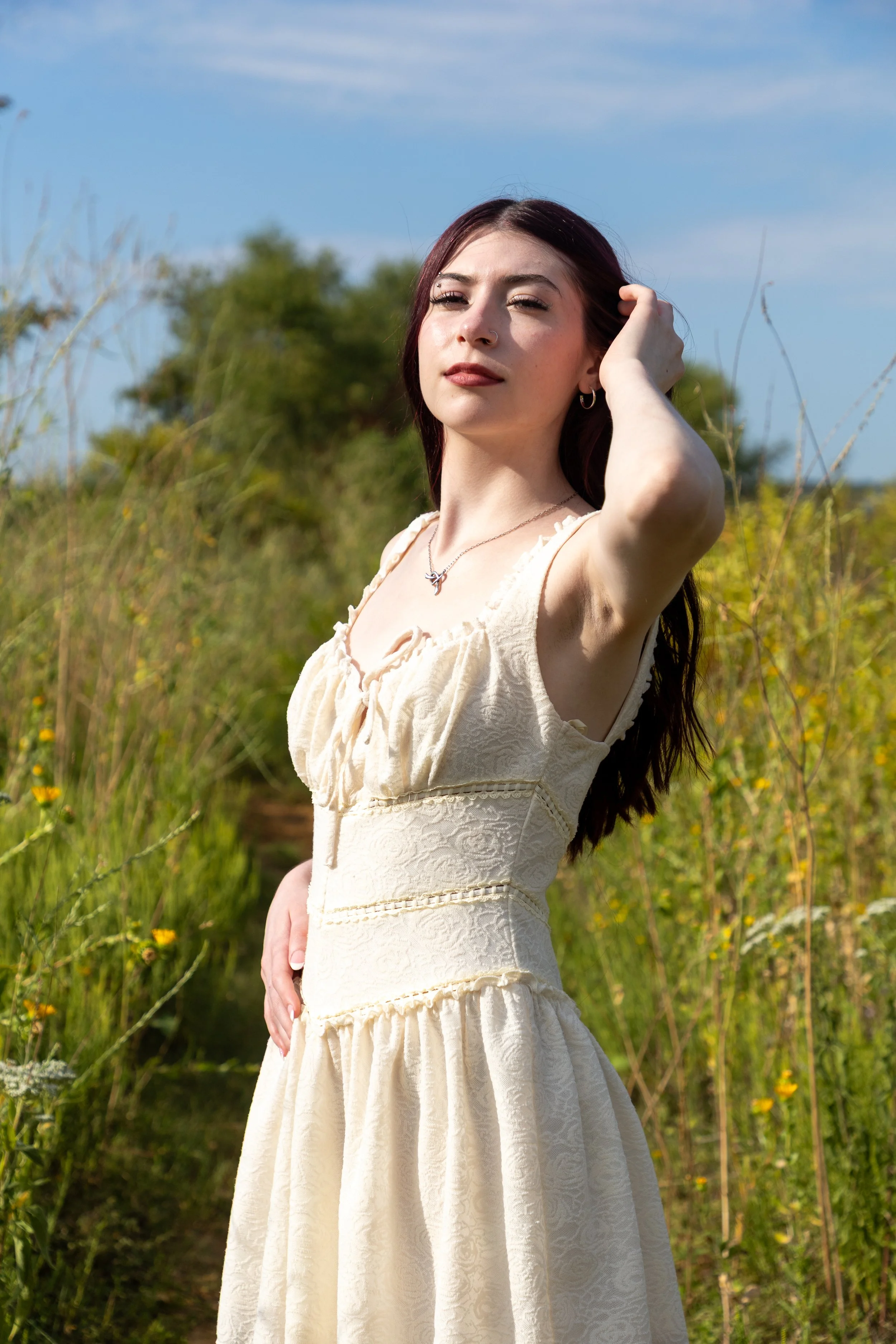 A young woman with dark hair in a cream-colored dress standing in a field of wildflowers under a blue sky.