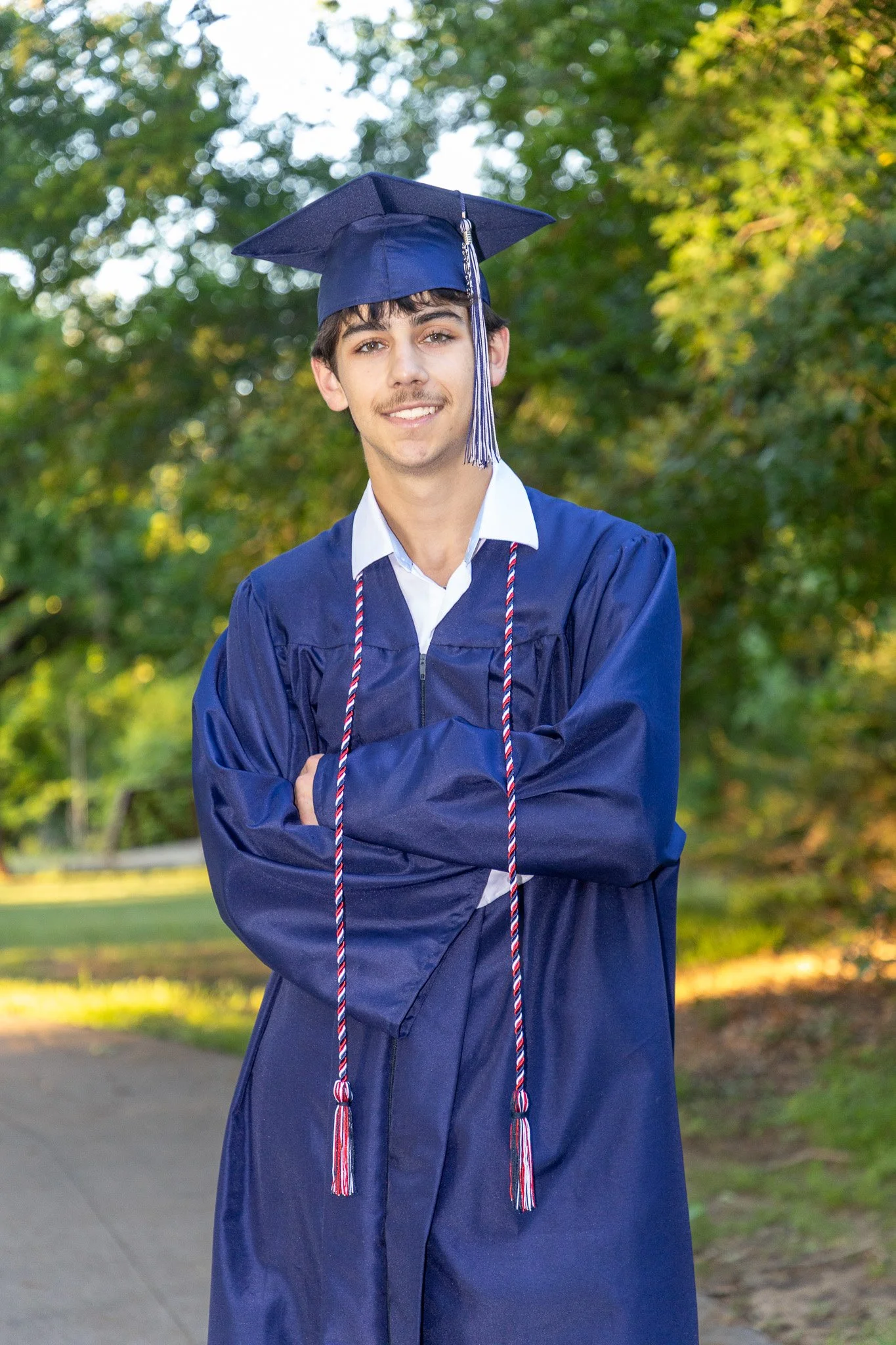 Young man in a graduation gown and cap standing outdoors in front of a tree, smiling with arms crossed.