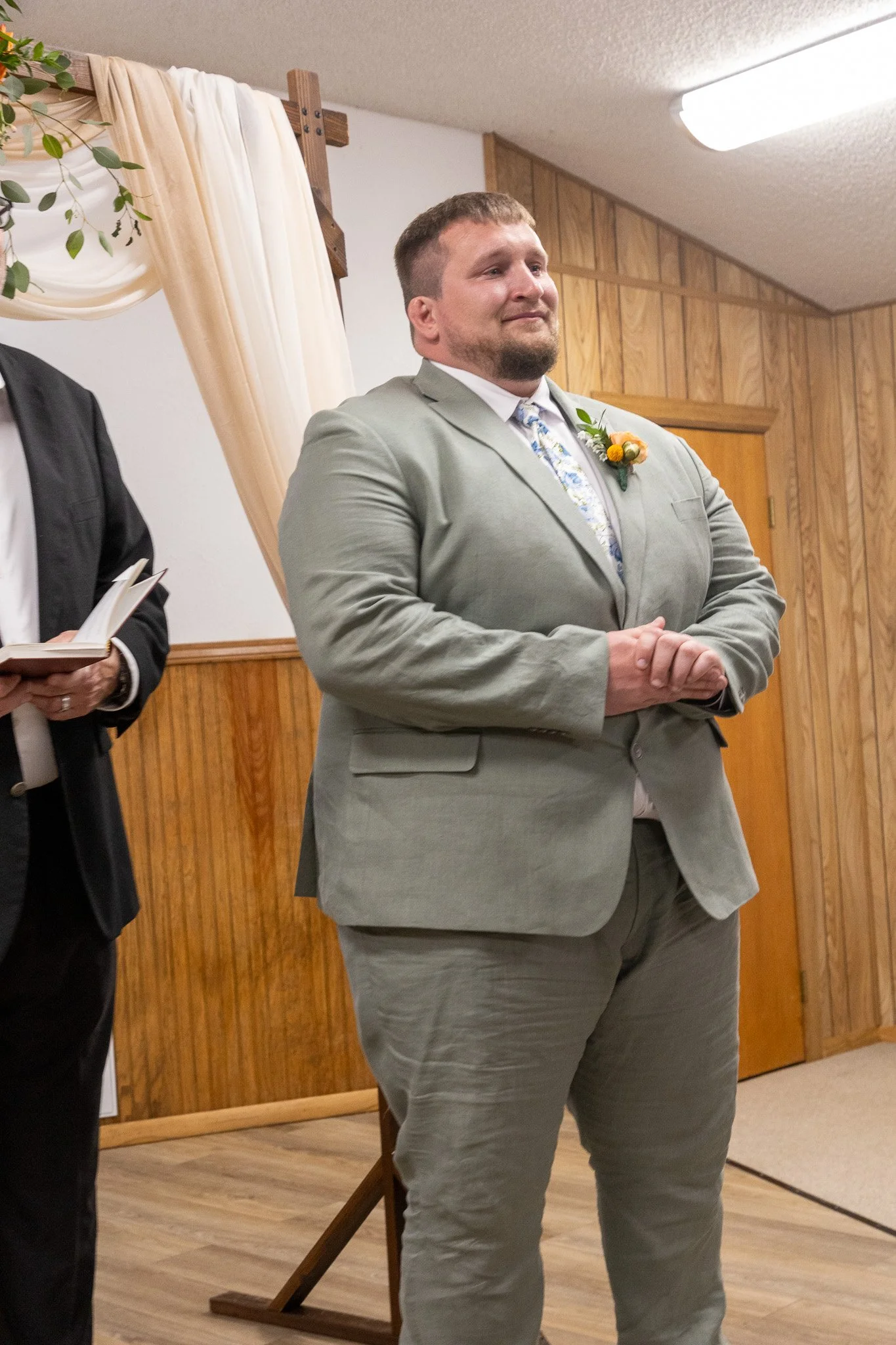 A man in a light gray suit is standing with a serious expression during a wedding ceremony, with a floral boutonniere on his left lapel and an officiant holding a book beside him.
