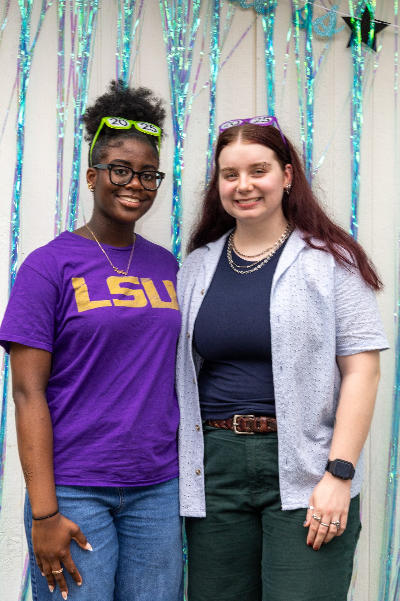 Two young women standing together, smiling, in front of a shiny, colorful party backdrop with tinsel and streamers.