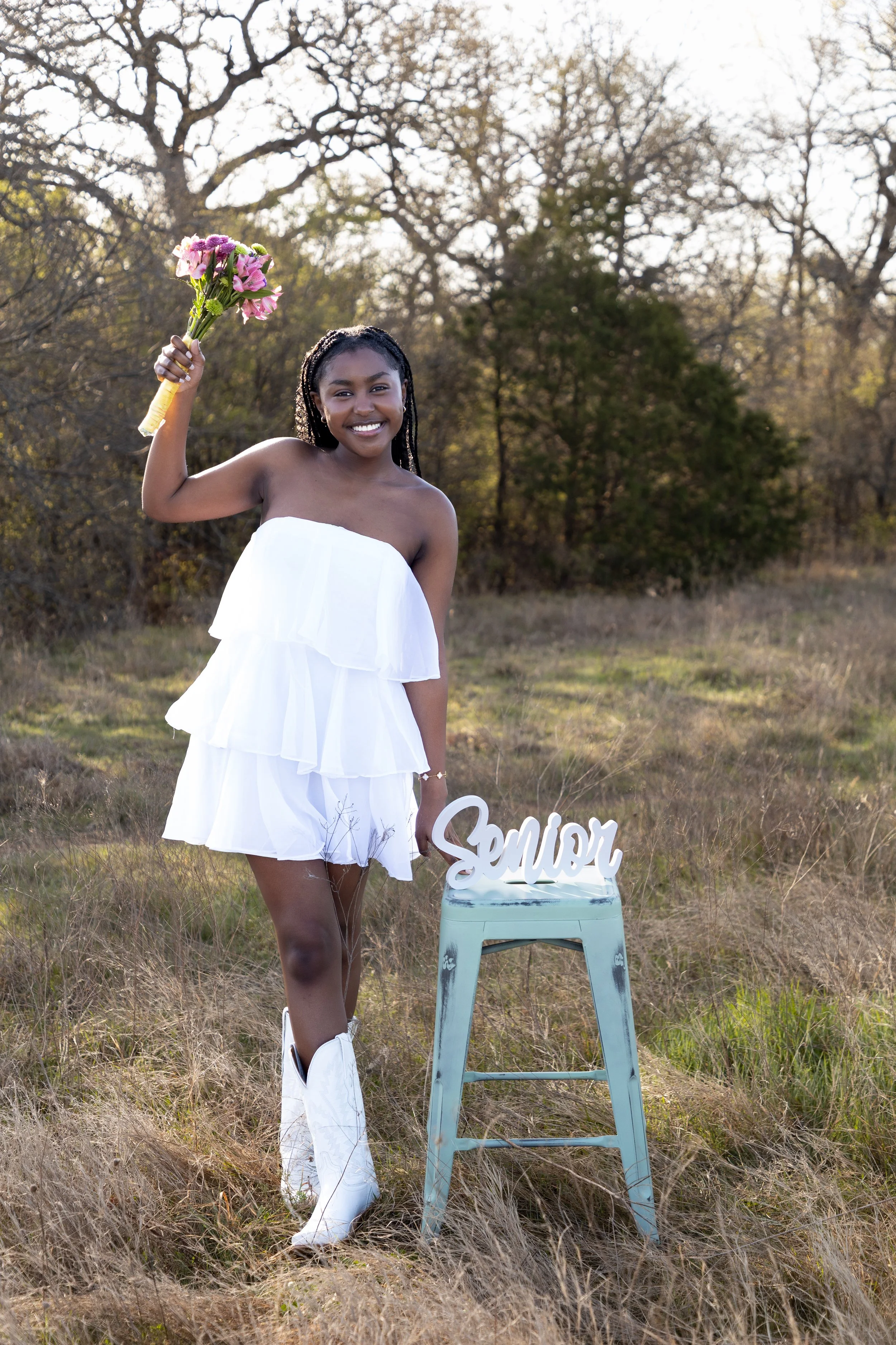 A young woman in a white strapless dress and white cowboy boots standing outdoors in a field with trees in the background, holding a bouquet of pink flowers above her head and smiling. Next to her is a small blue stool with a decorative sign that rea
